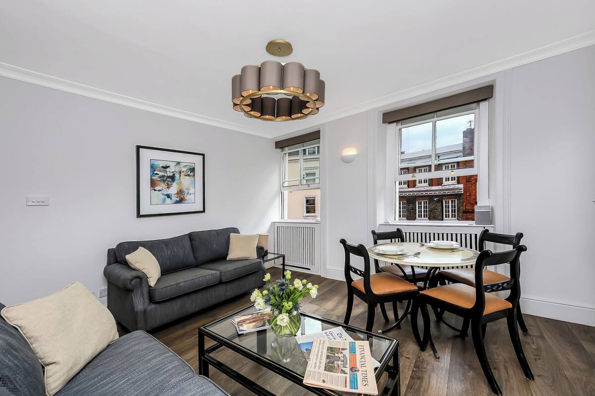 Een moderne woonkamer met een grijze bank, een salontafel met een vaas en krant, en een eethoek met een ronde tafel en stoelen.