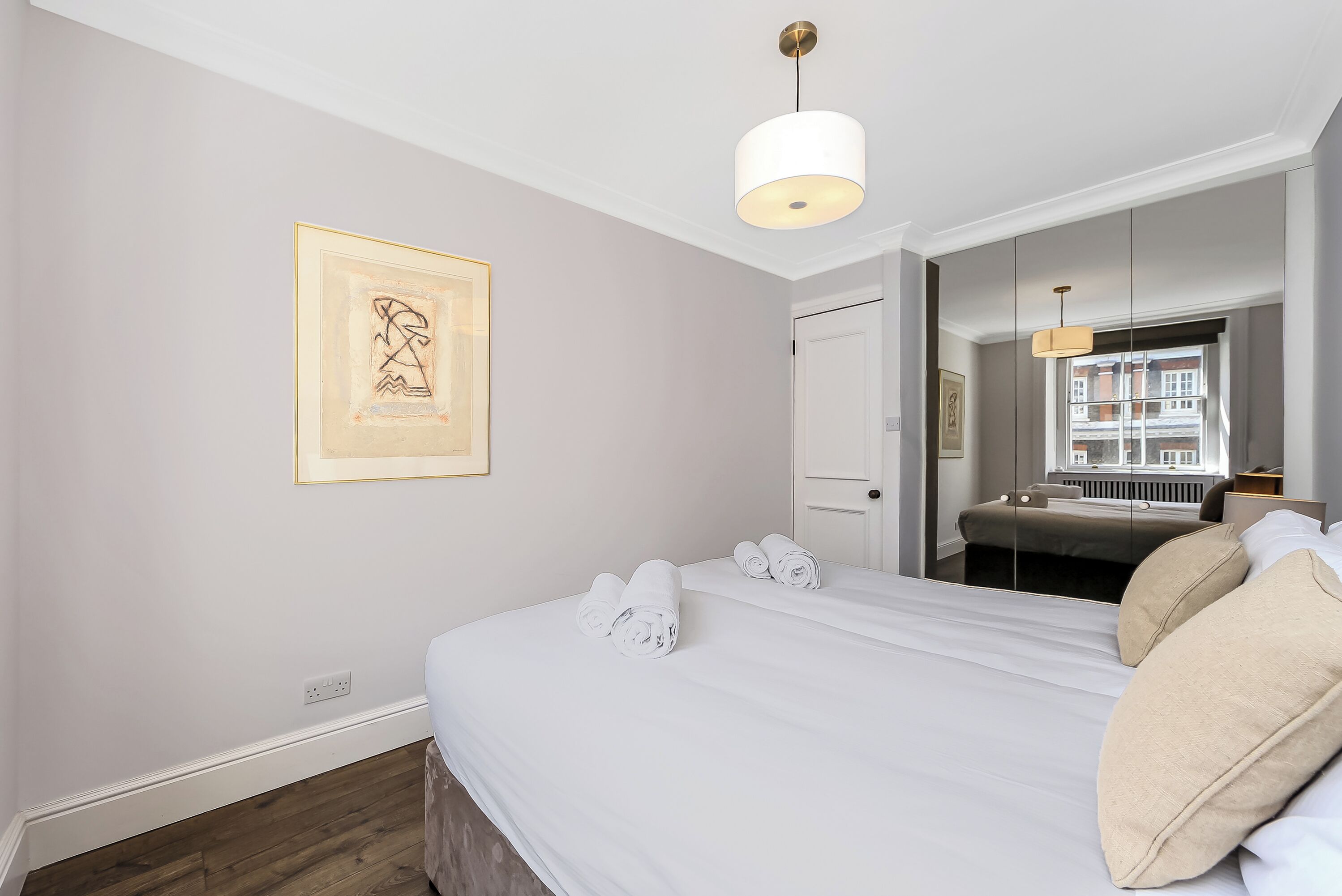 A neatly made bed with rolled towels, a framed artwork on the wall, and a window with a view of buildings.