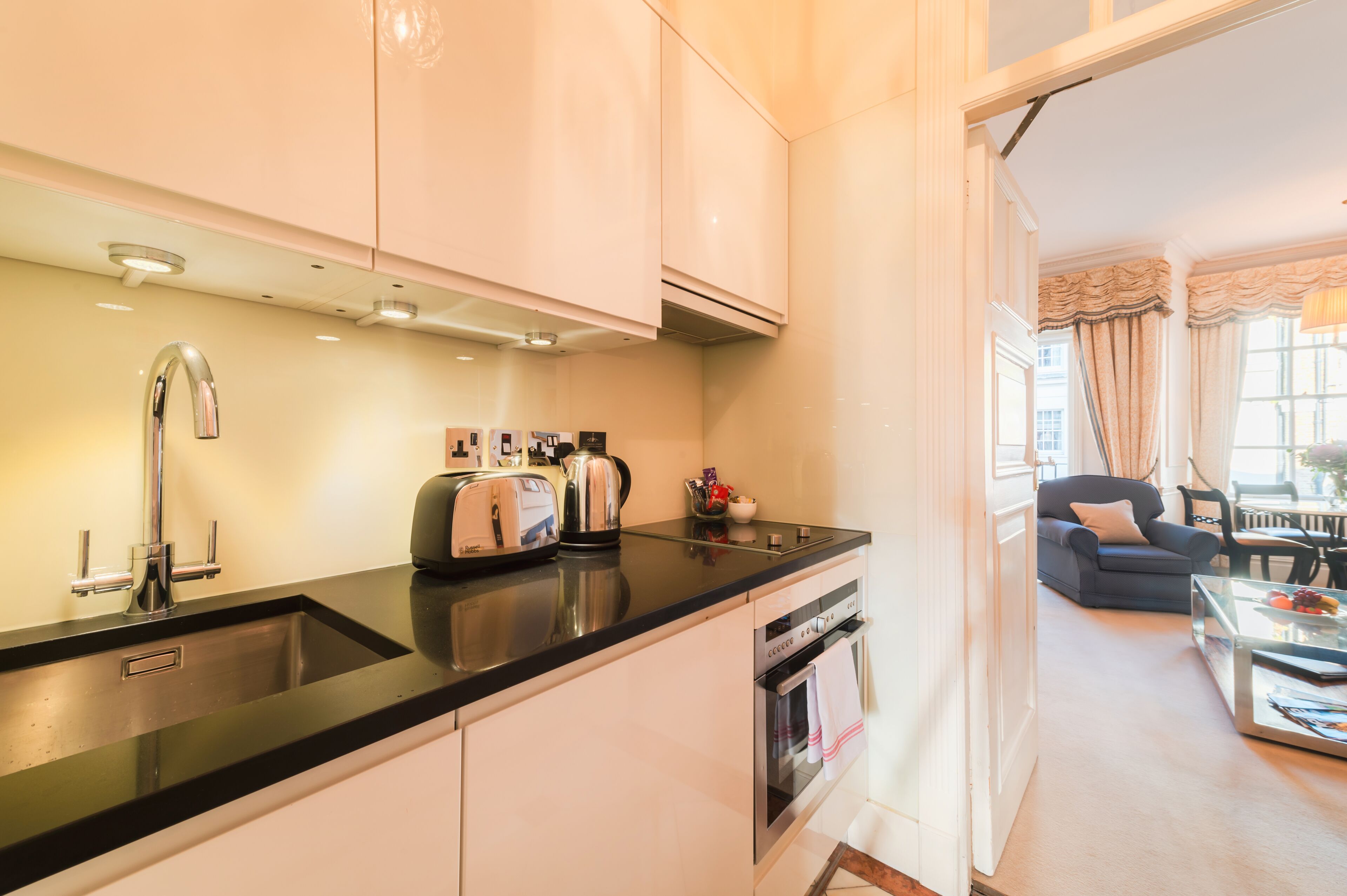 Uma cozinha moderna com bancada preta, eletrodomésticos de aço inoxidável e vista para uma sala de estar com sofá e mesa de centro.