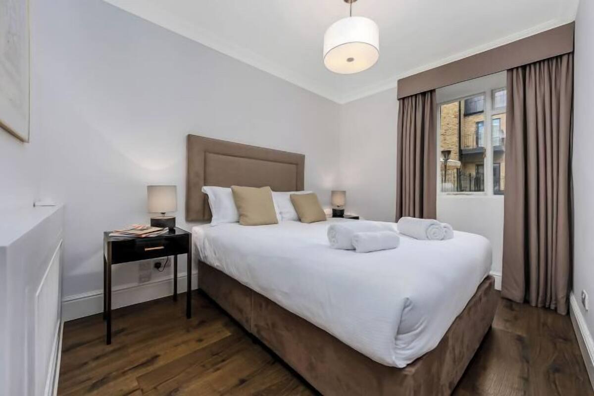 Una habitación de hotel con una cama grande, dos mesitas de noche, una lámpara colgante y una ventana con cortinas.