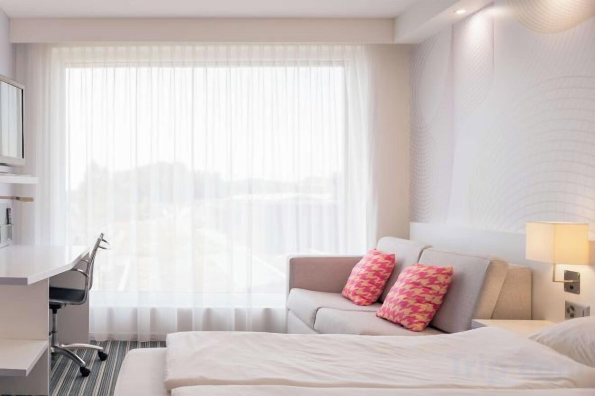 A hotel room with a bed, a desk, a chair, and a sofa with pillows.
