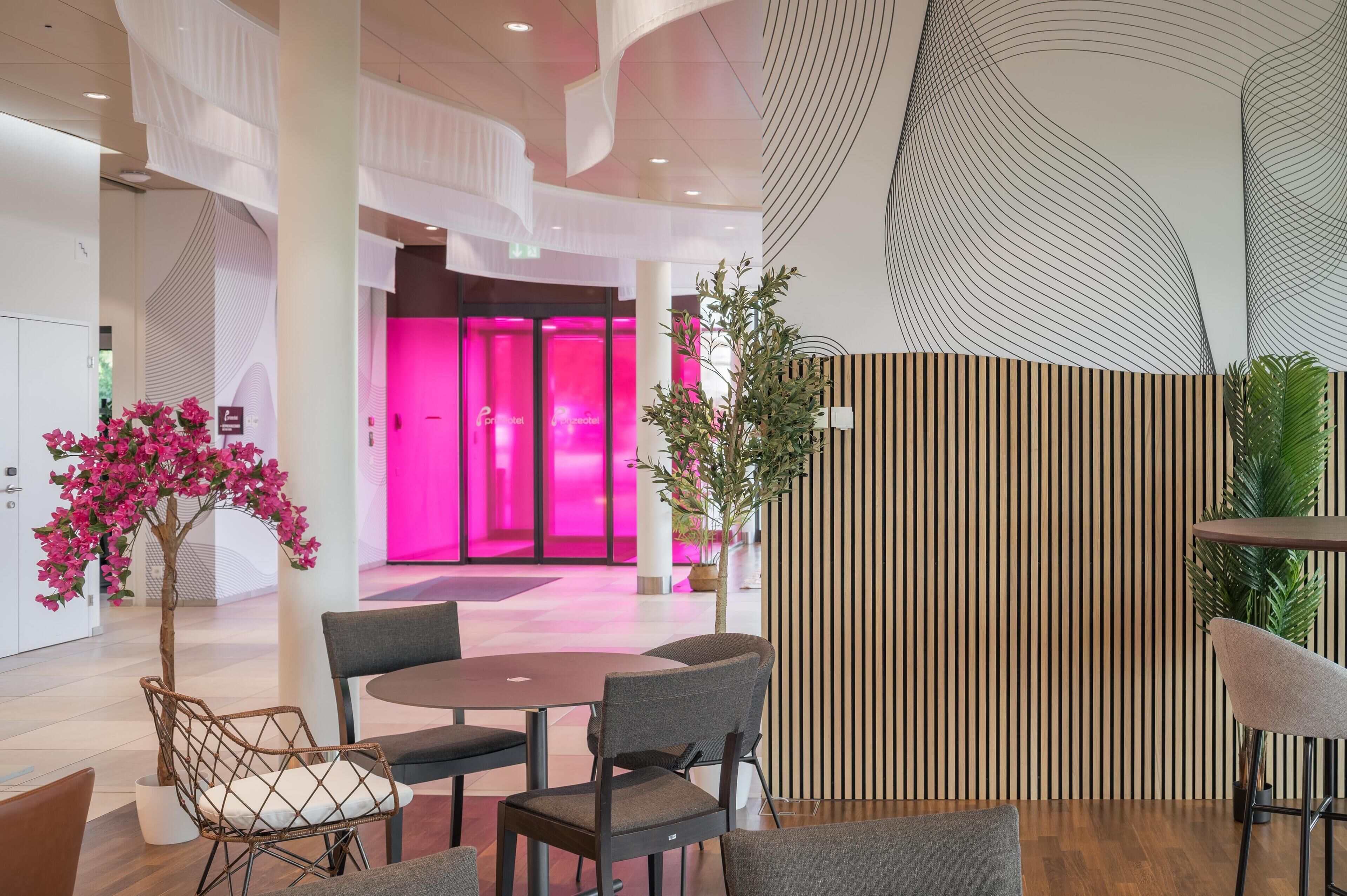 Eine moderne Lobby mit einem runden Tisch und Stühlen, einem Blumentopf und einem im Hintergrund leuchtend rosa erhellten Raum.