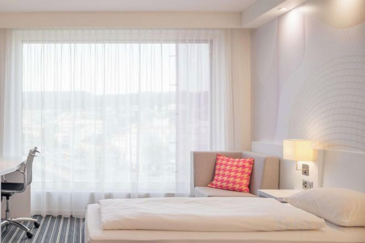 A hotel room with a bed, a desk, a chair, a lamp, and a window with sheer curtains.