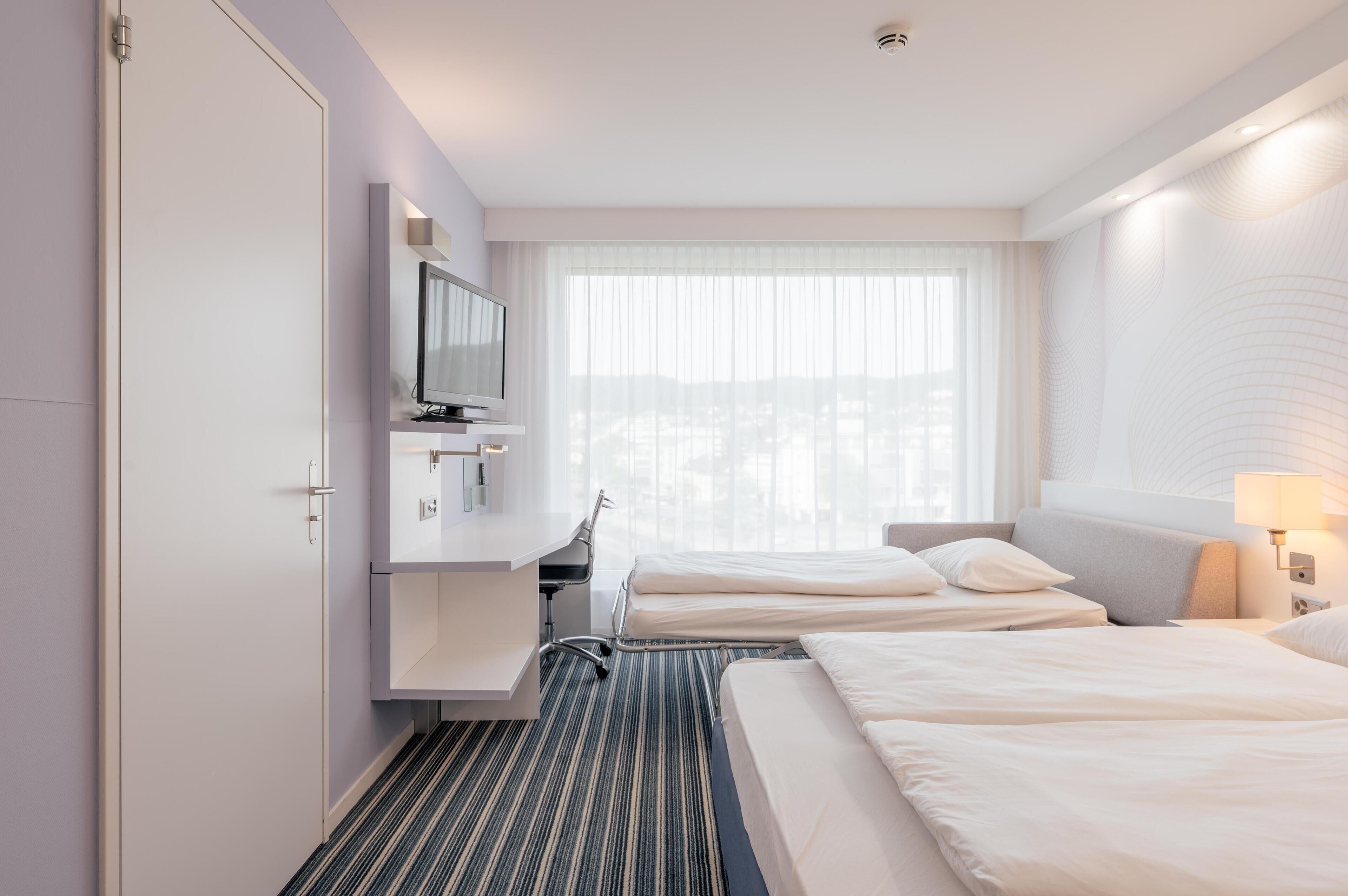 Quarto de hotel com duas camas, uma escrivaninha com televisão, uma janela com cortinas translúcidas e uma luminária fixada na parede.