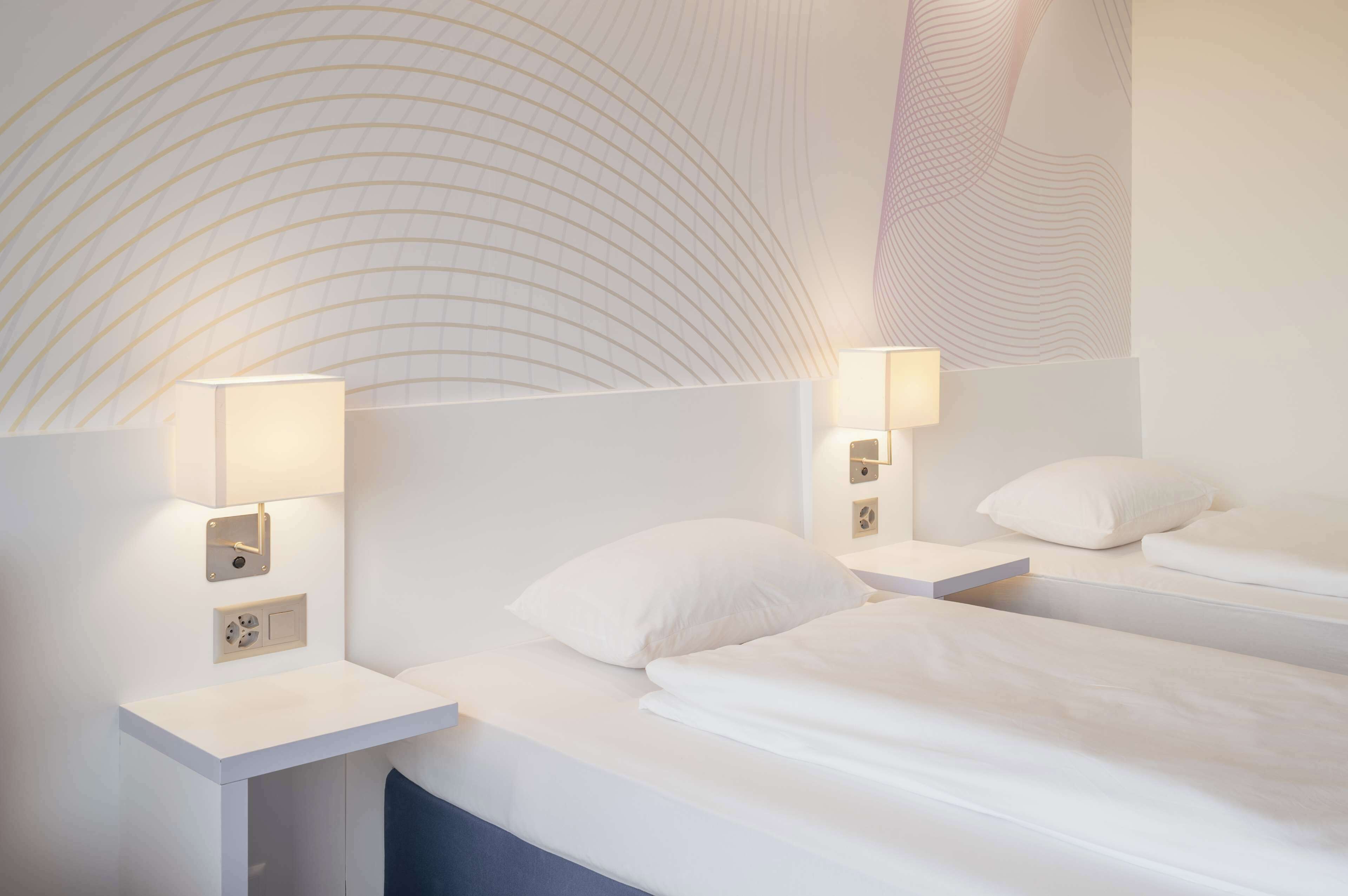 Ein Hotelzimmer mit zwei Betten, jeweils mit Nachttisch und Lampe, sowie einem großen Wandbild.