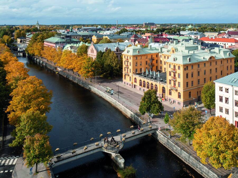 A river with a pedestrian bridge, surrounded by autumn-colored trees and historic buildings.