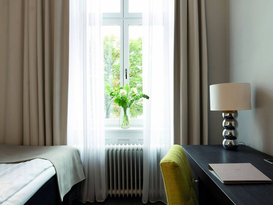 Habitación de hotel con cama, escritorio con lámpara, silla y ventana con cortinas.