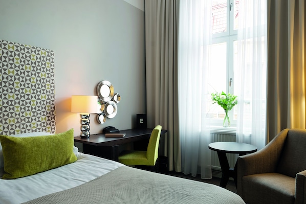 Quarto de hotel com cama, escrivaninha com luminária, cadeira, mesinha e janela com cortinas.