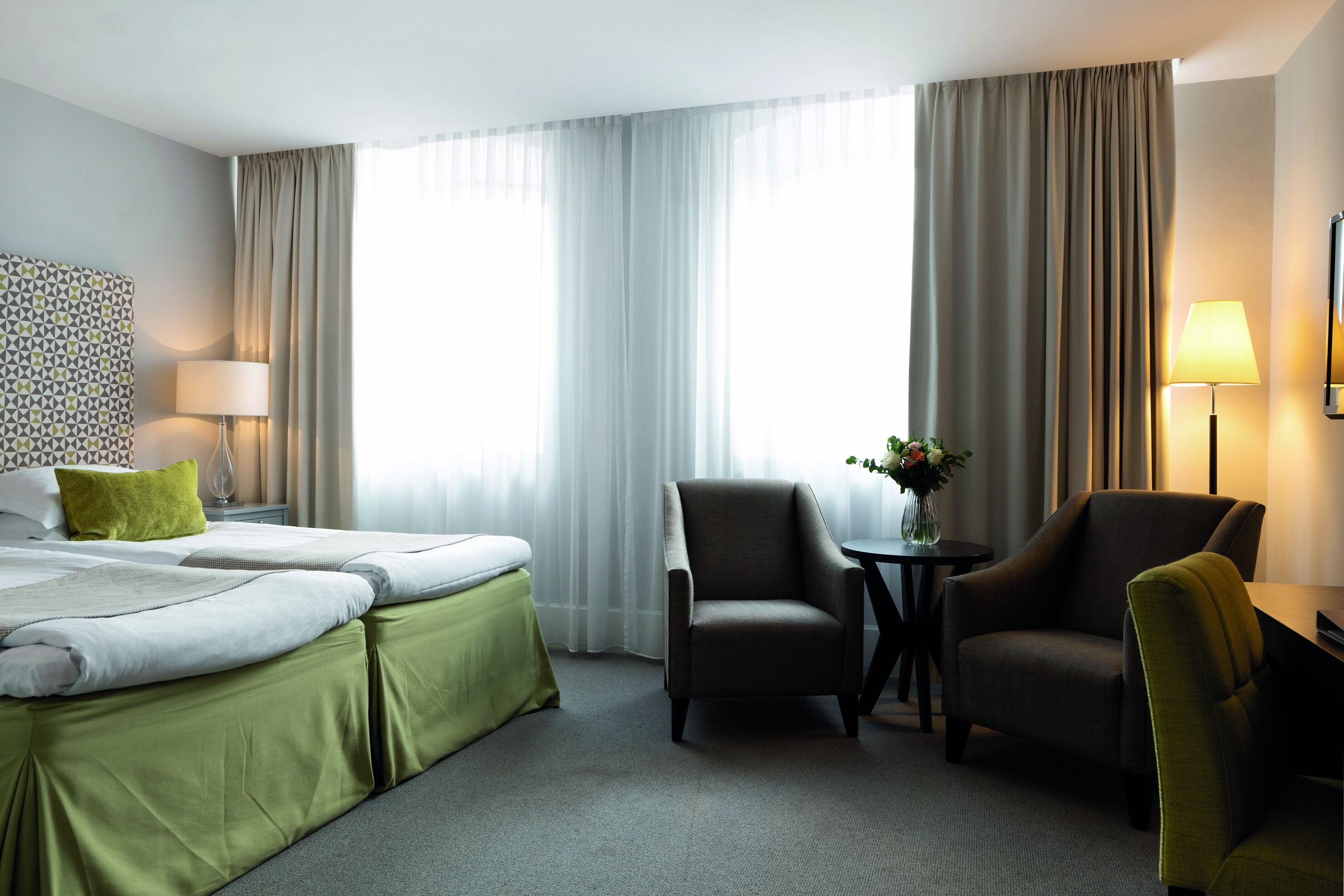 A hotel room with a bed, two chairs, a small table with a vase, and a lamp.