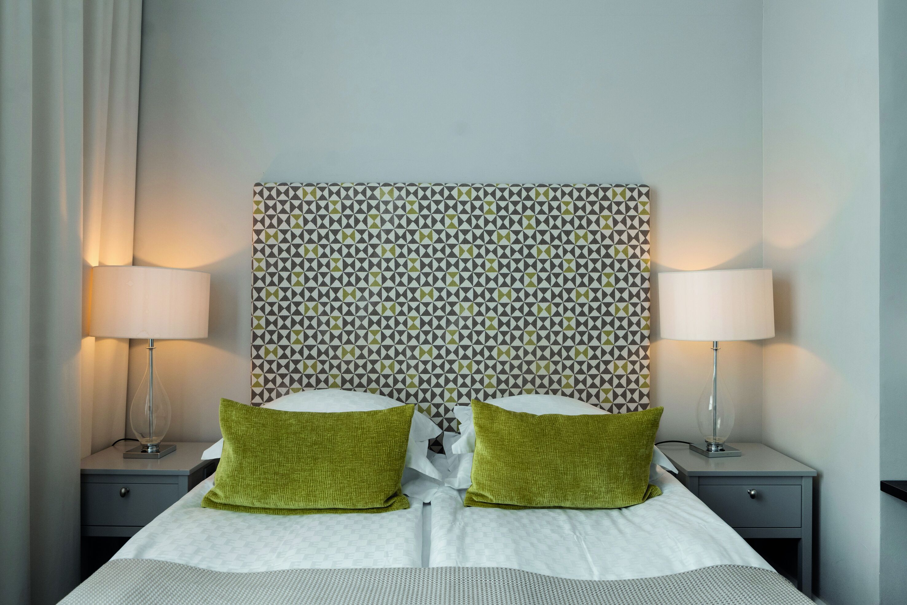 Una cama bien tendida con dos almohadas verdes, dos lámparas de noche y un cabecero estampado.