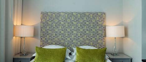 Una cama bien tendida con dos almohadas verdes, dos lámparas de noche y un cabecero estampado.