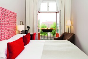 A hotel room with a large bed, red pillows, a desk with a lamp, and a view of a balcony with greenery. - Elite Grand Hotel Gävle (Gävle)