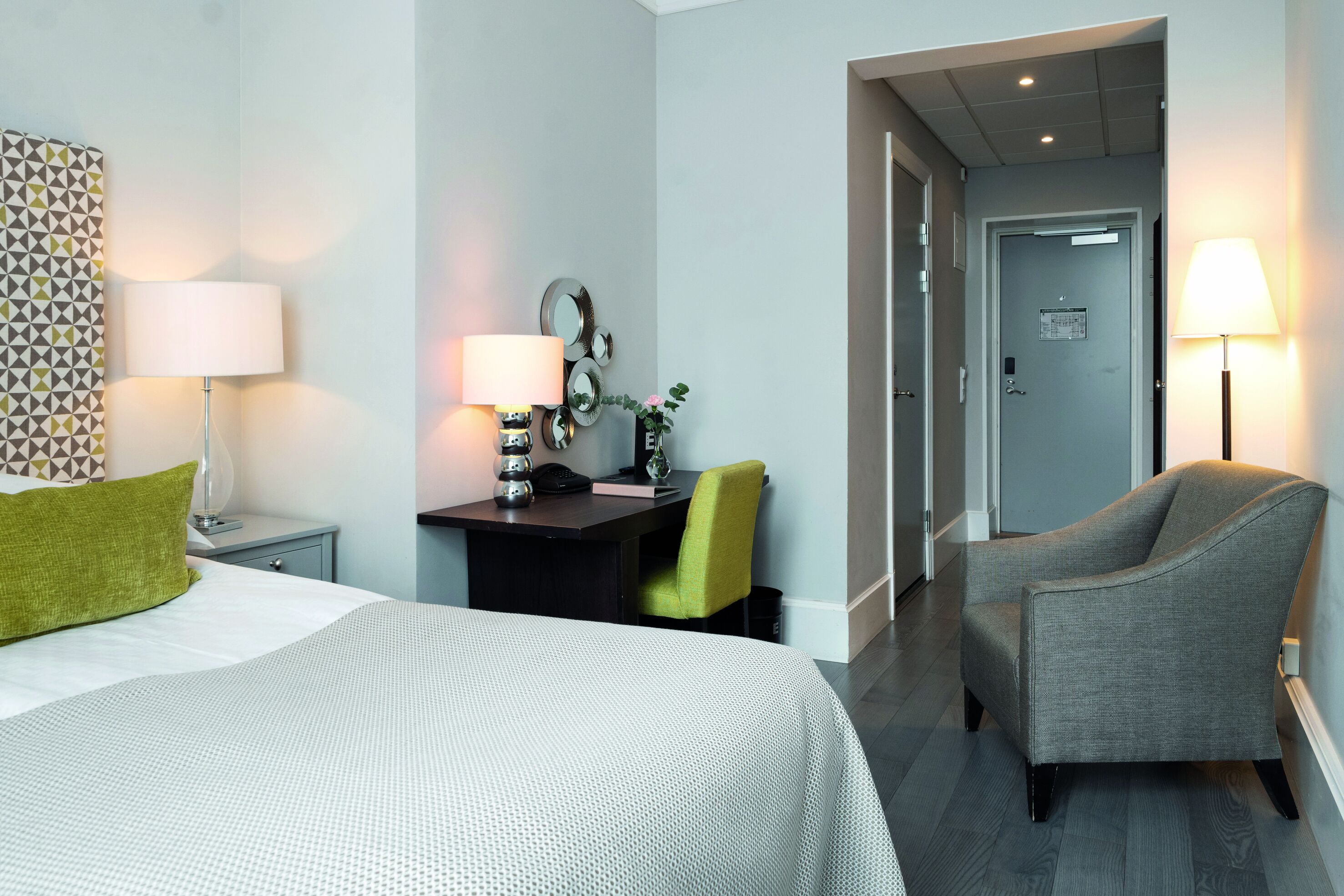 A hotel room with a bed, a desk with a lamp, a chair, and a door.