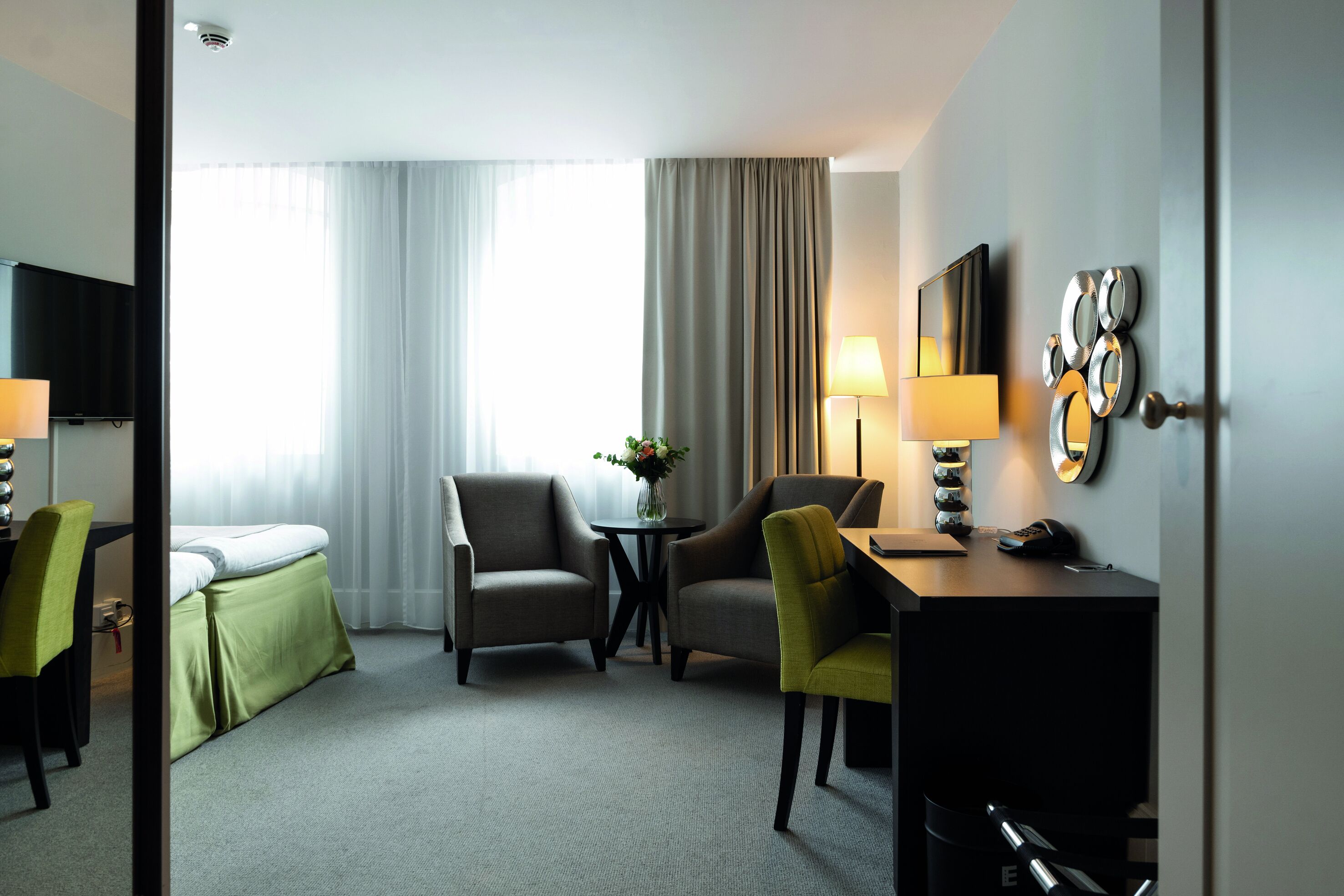 Une chambre d’hôtel avec un lit, deux fauteuils, un bureau avec une lampe, une petite table avec un vase et un miroir au mur.