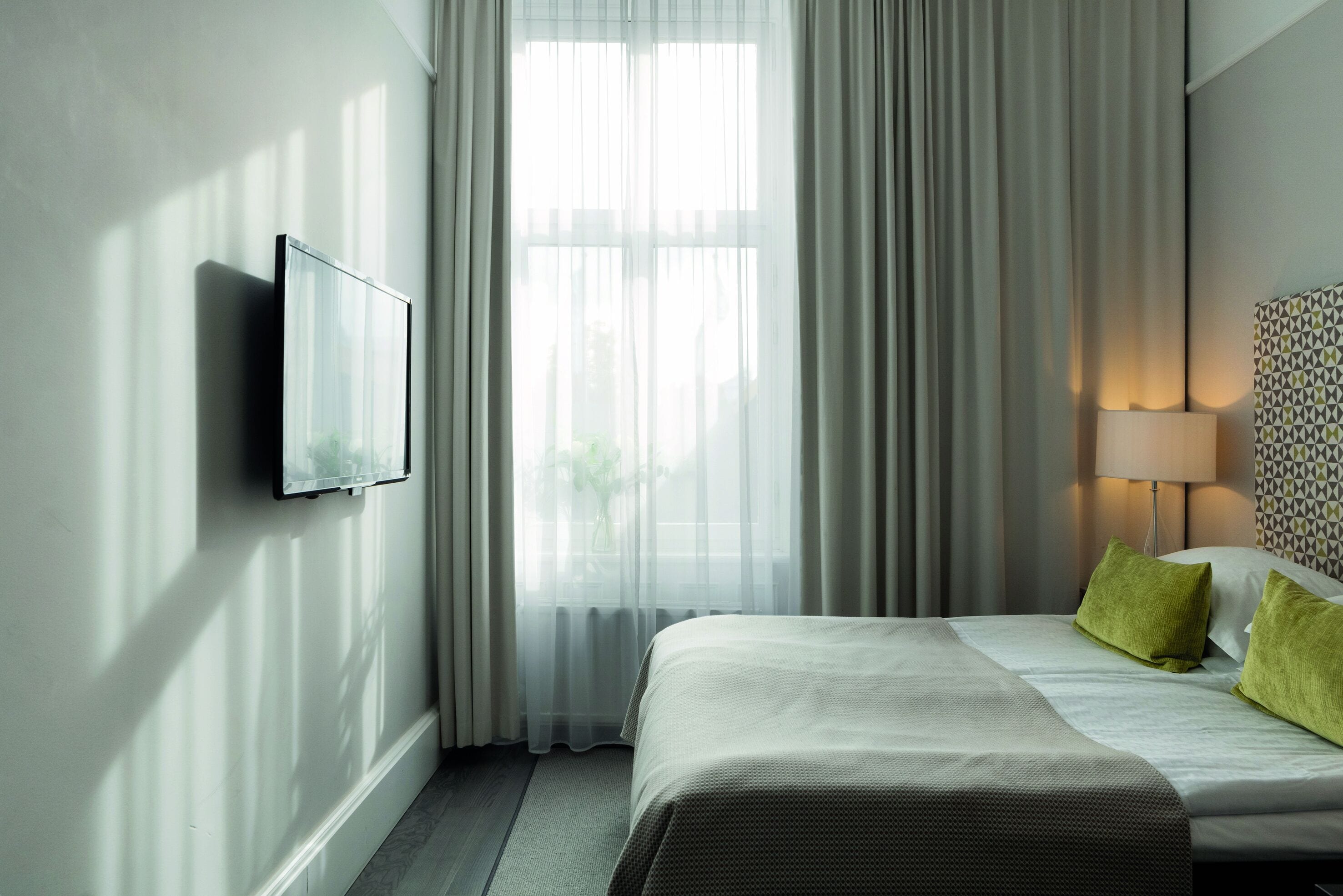 A hotel room with a bed, a TV mounted on the wall, a lamp, and a window with curtains.