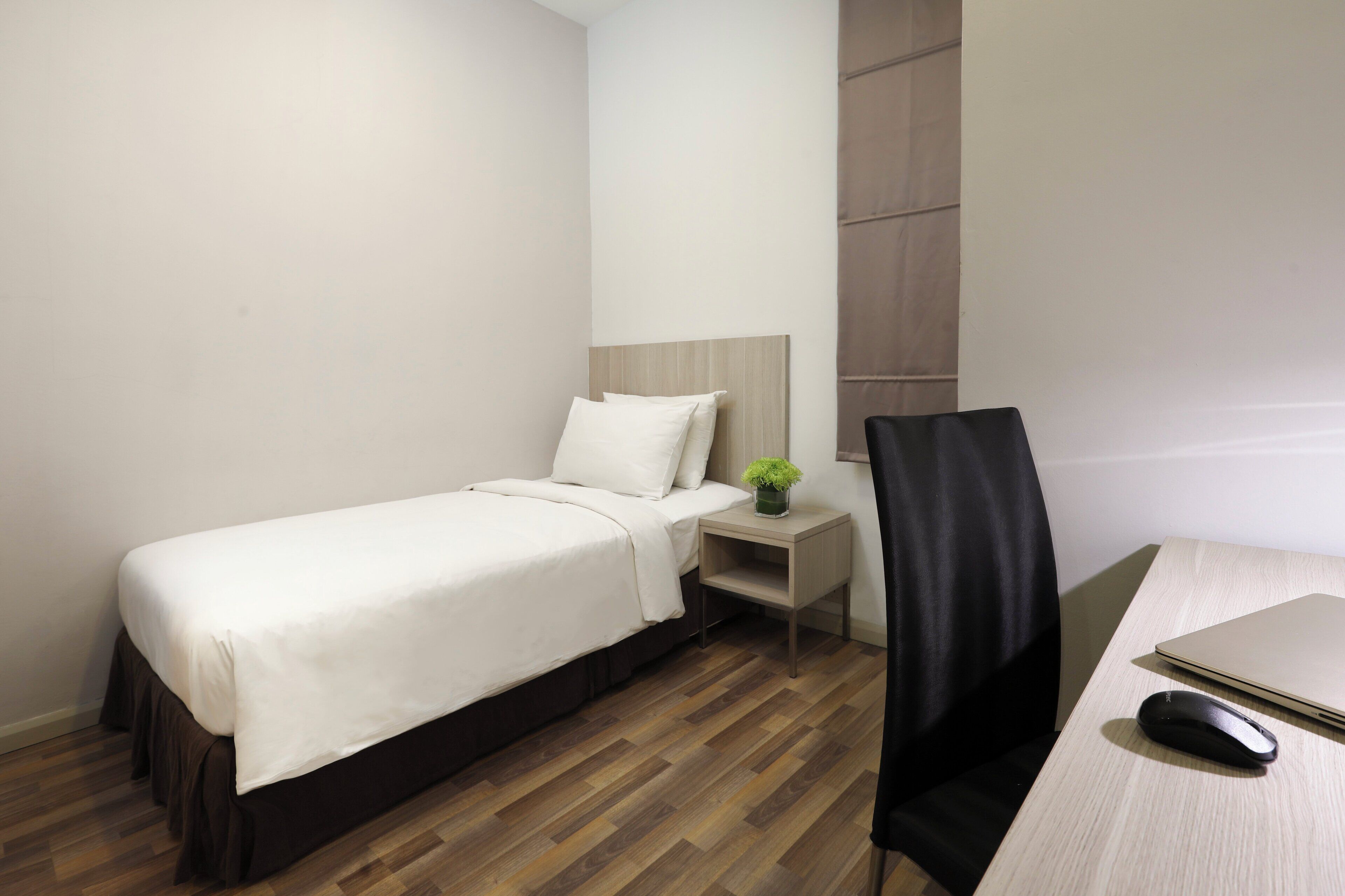 A hotel room with a bed, a small bedside table with a plant, a desk with a laptop and mouse, and a chair.