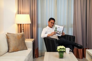 Un hombre sentado en una butaca de cuero negro leyendo una revista en una habitación luminosa, con una lámpara, un sofá y una mesa de centro con un florero.