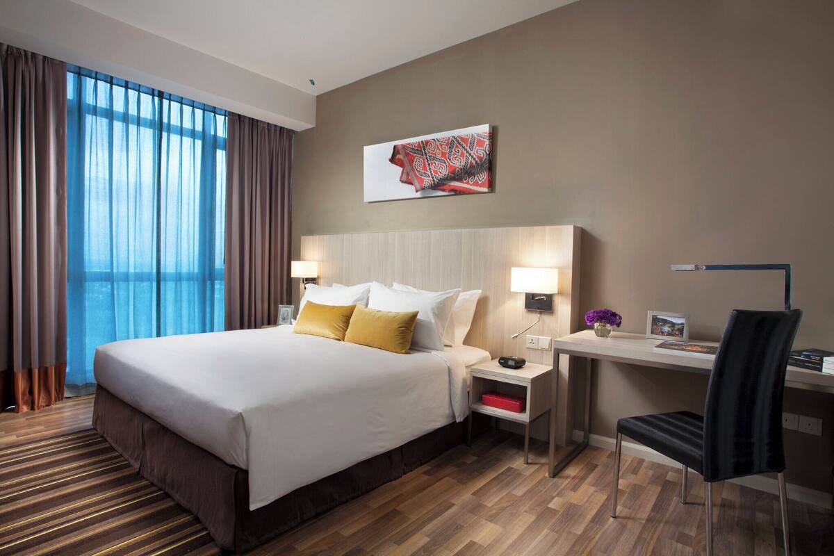 A hotel room with a large bed, two bedside tables with lamps, a desk with a chair, and a large window with curtains.