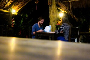 Un couple est assis à une table en bois dans une pièce à la lumière tamisée, dont le toit est recouvert de chaume, et qui est entourée de plantes tropicales.