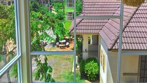 A view through a window of a building with a tiled roof, a water tower in the background, and a green area with trees and a bench. - Green Home Resort (Surat Thani)