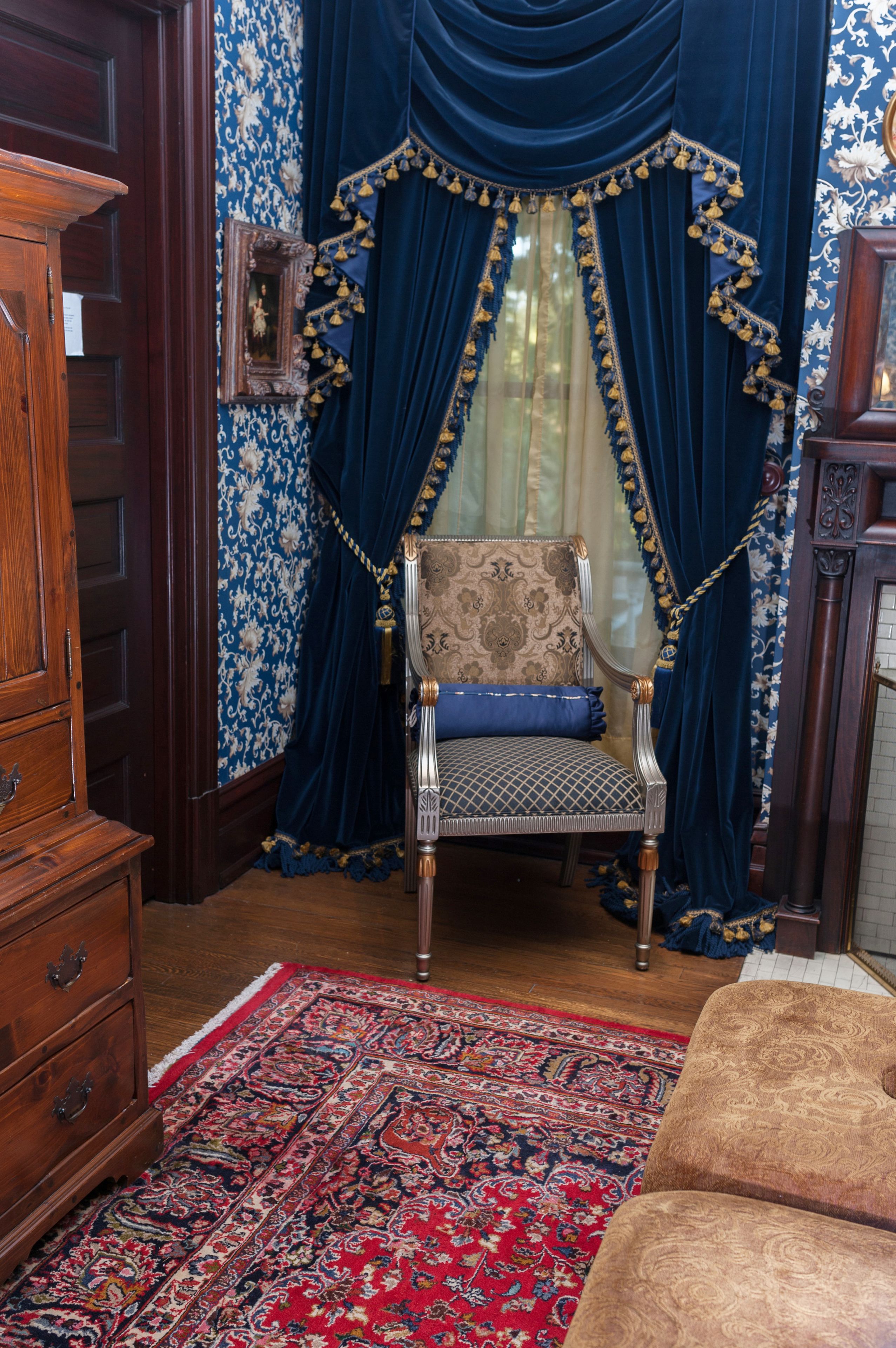 A room with a patterned rug, a wooden dresser, a chair with a blue cushion, and blue curtains with tassels.