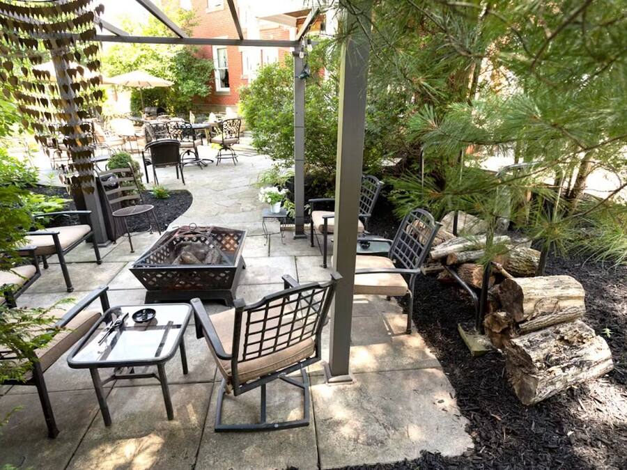 An outdoor patio area with seating arrangements, including tables and chairs, surrounded by greenery and a fire pit.