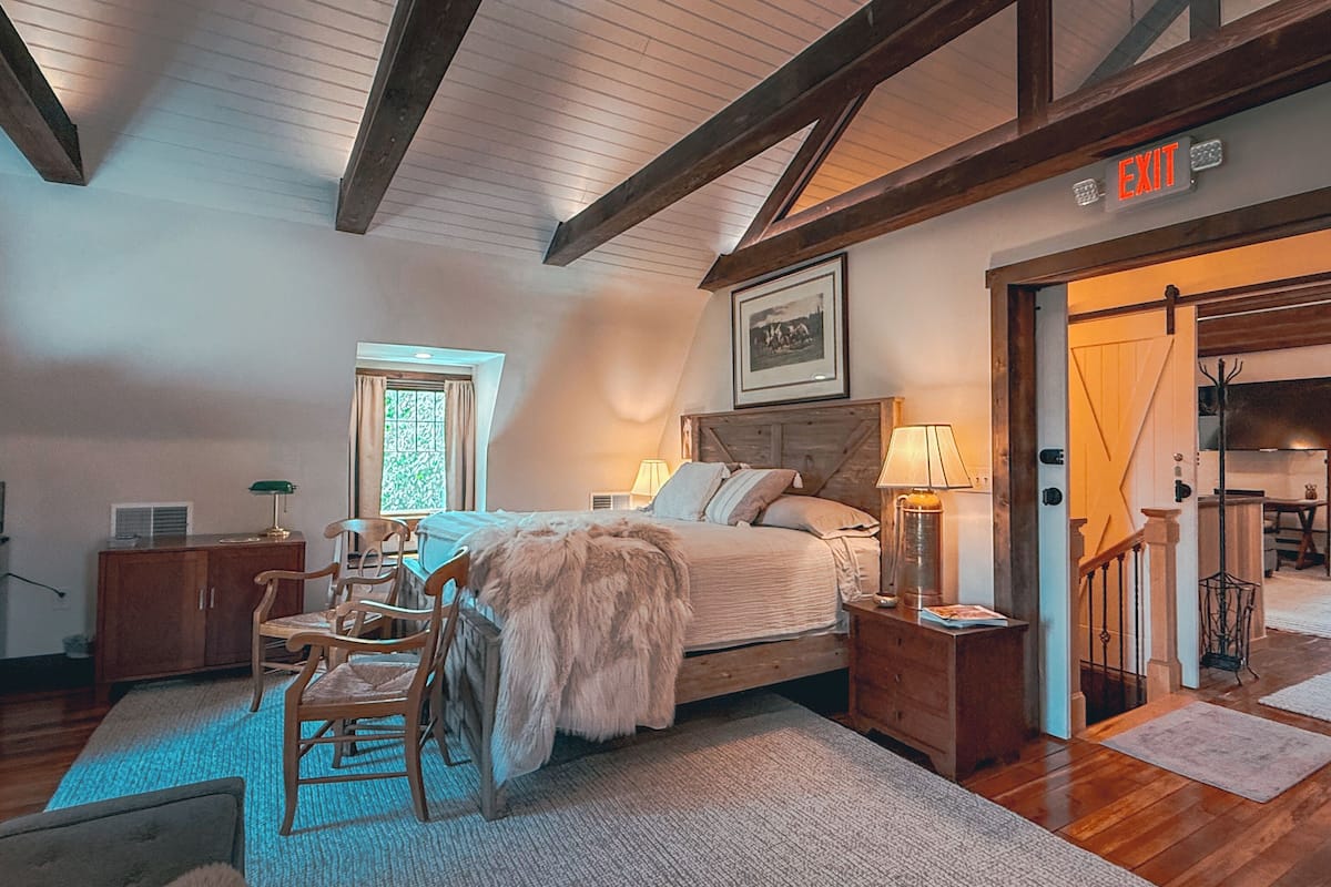 Quarto com cama grande, cabeceira de madeira, mesa de cabeceira, cômoda, cadeira, janela e placa de saída.
