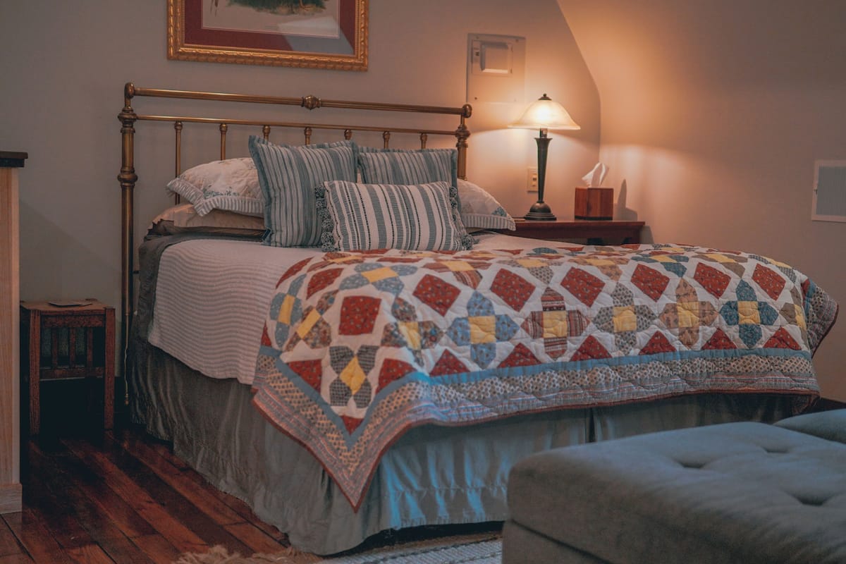 Uma cama bem arrumada com um edredom, um abajur e uma pequena mesa de cabeceira de madeira.