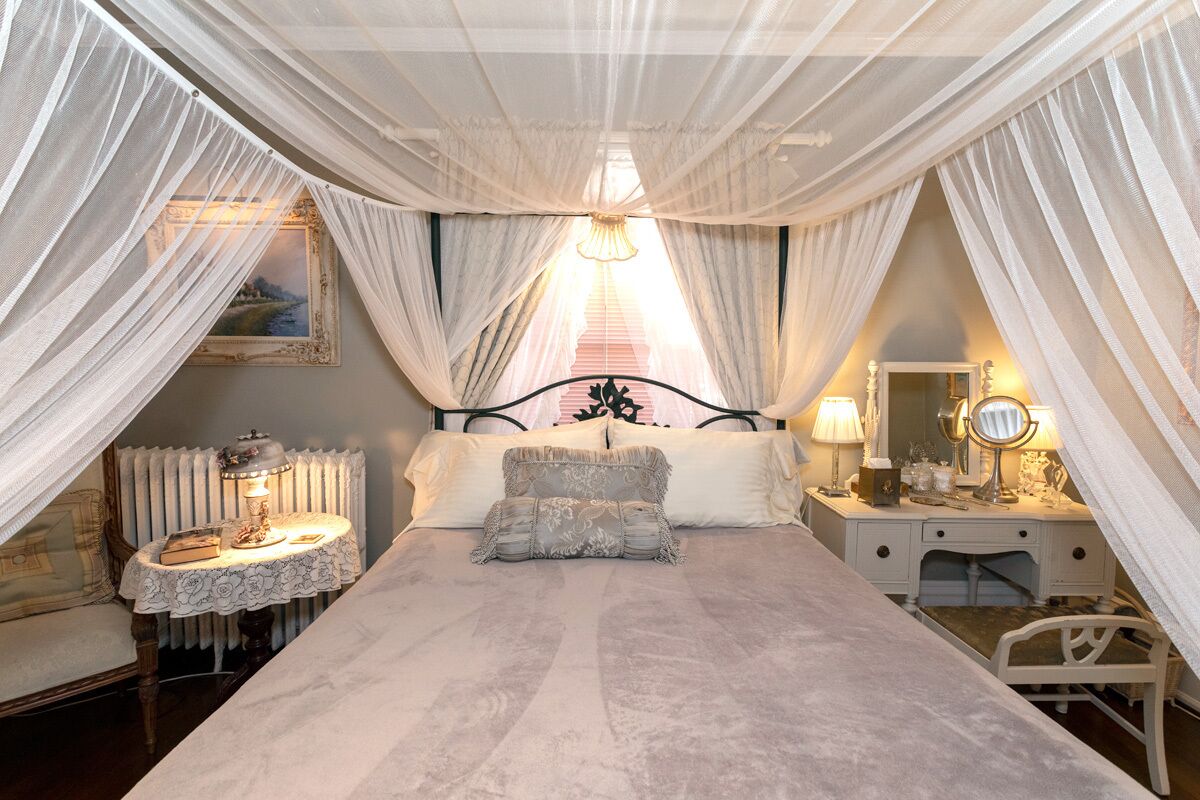 A bedroom with a large bed, a bedside table with a lamp, a dresser with a mirror, and sheer curtains.