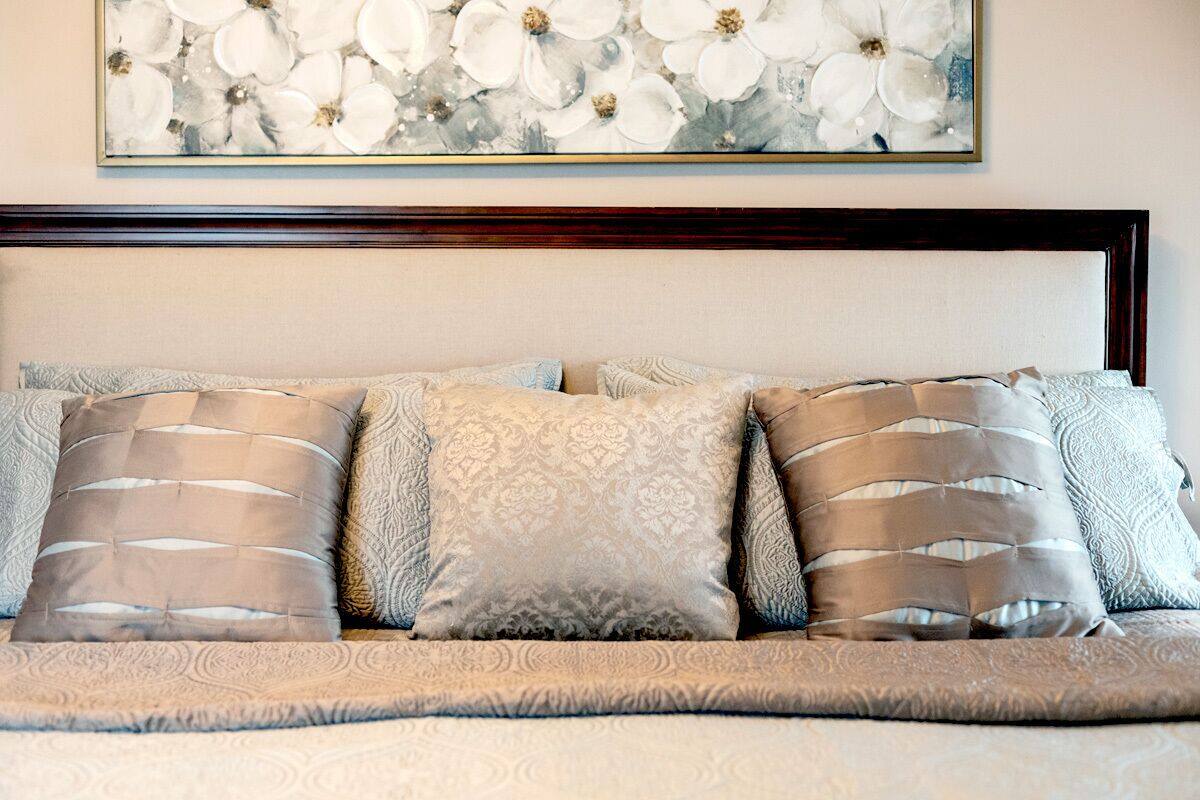 Cama bem arrumada com colcha estampada e várias almofadas, cabeceira de madeira e uma obra de arte floral acima.