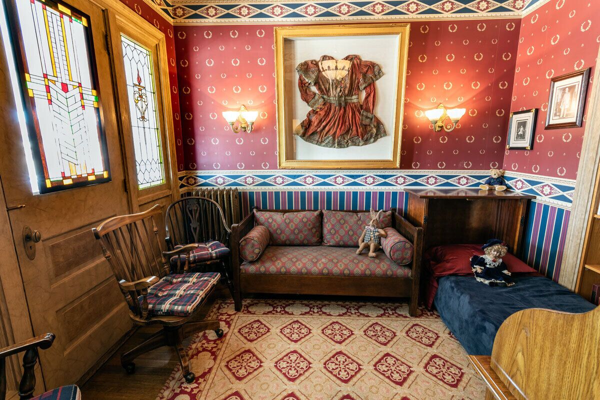 Une pièce avec un plancher en bois, un canapé agrémenté de coussins à motifs, un petit lit, une chaise en bois, un tableau encadré accroché au mur et un tapis à motifs.