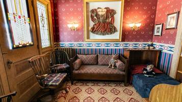 Une pièce avec un plancher en bois, un canapé agrémenté de coussins à motifs, un petit lit, une chaise en bois, un tableau encadré accroché au mur et un tapis à motifs.