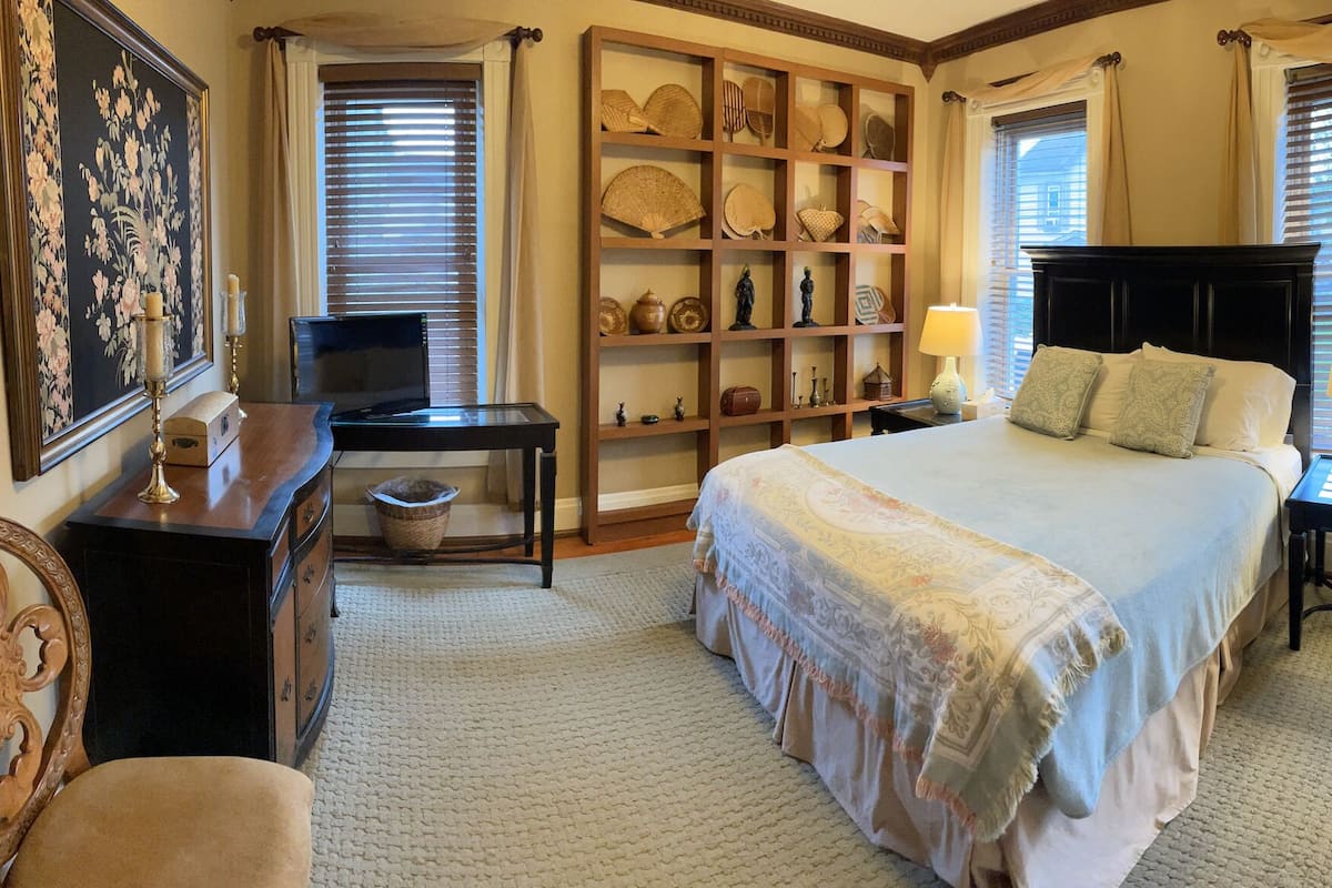 Quarto com cama grande, mesa de cabeceira de madeira, escrivaninha com computador, estante de livros e espelho.