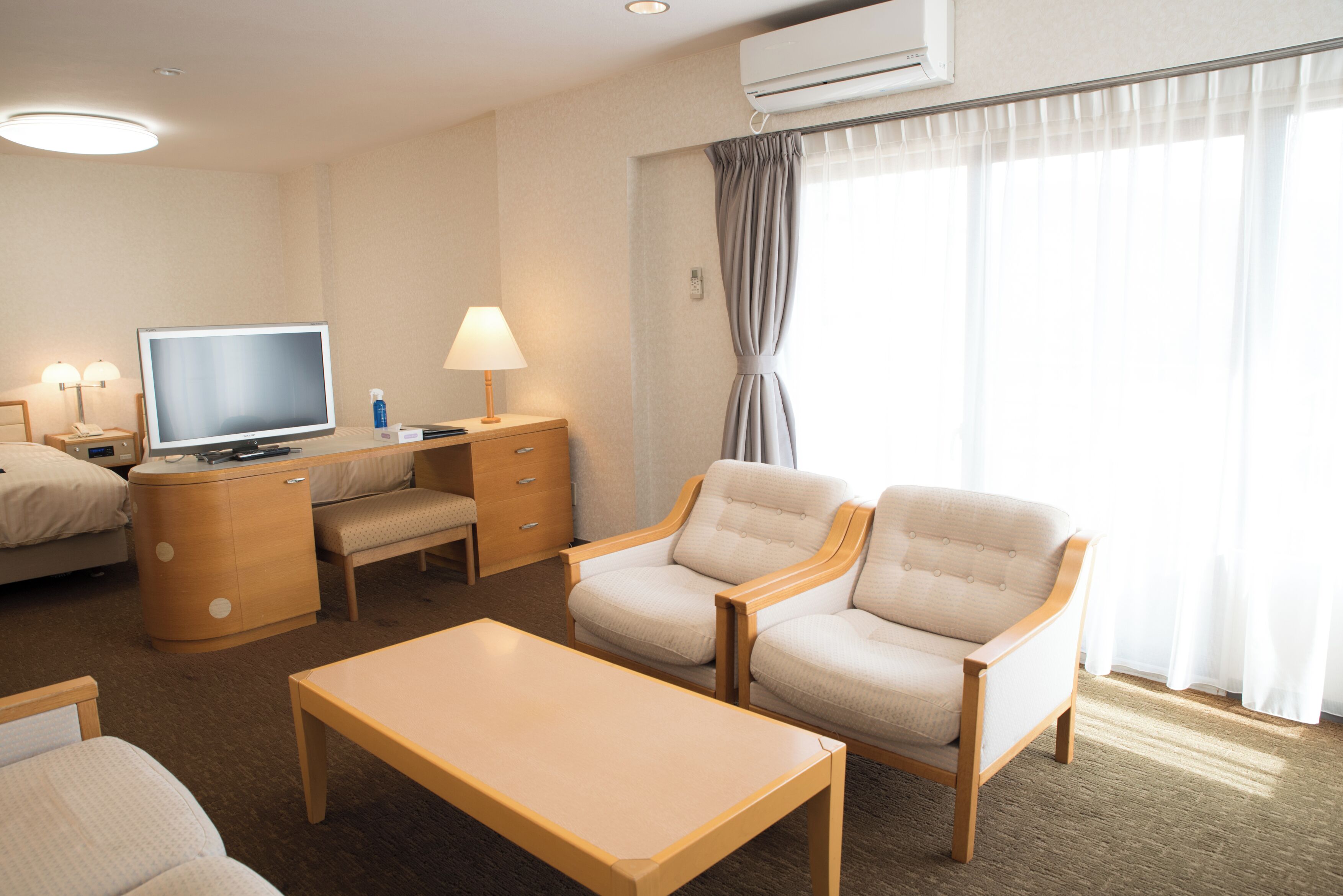 A hotel room with a bed, a desk with a computer, two armchairs, a coffee table, and a large window with curtains.