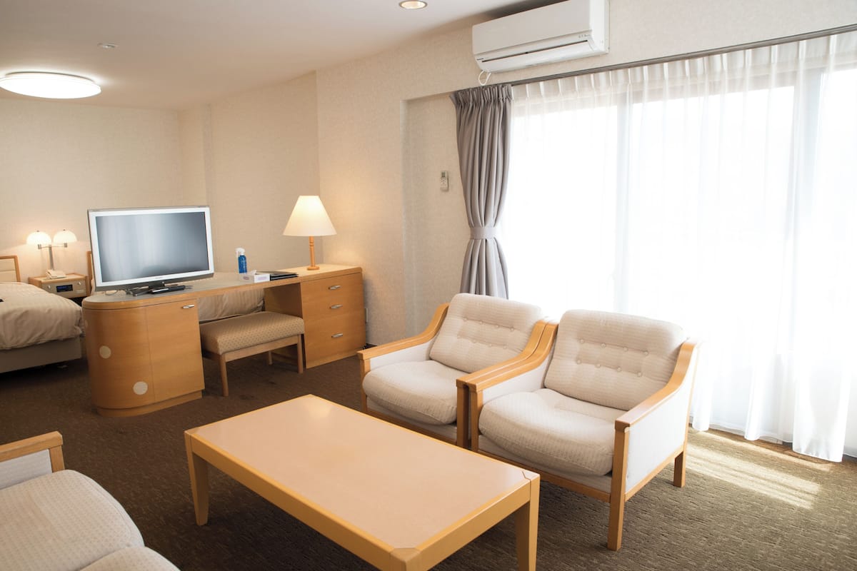 A hotel room with a bed, a desk with a computer, two armchairs, a coffee table, and a large window with curtains.