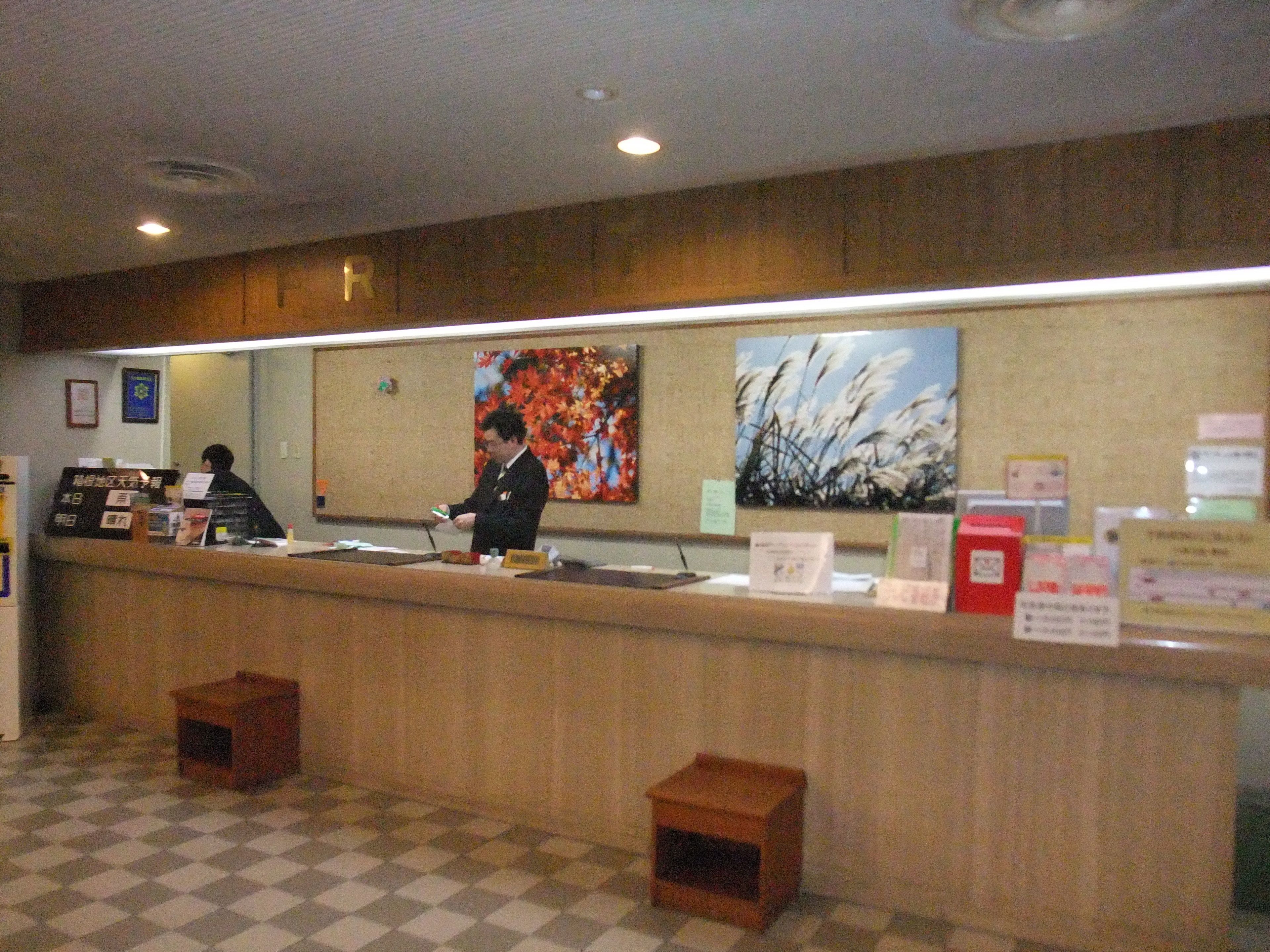 A reception area with a wooden counter, a check-in desk, and a check-out counter. There are two small wooden stools and a large painting on the wall.