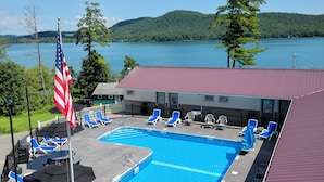 A swimming pool with lounge chairs and umbrellas, an American flag, and a view of a lake and forested hills. - Lake 'N Pines Motel (Cooperstown)