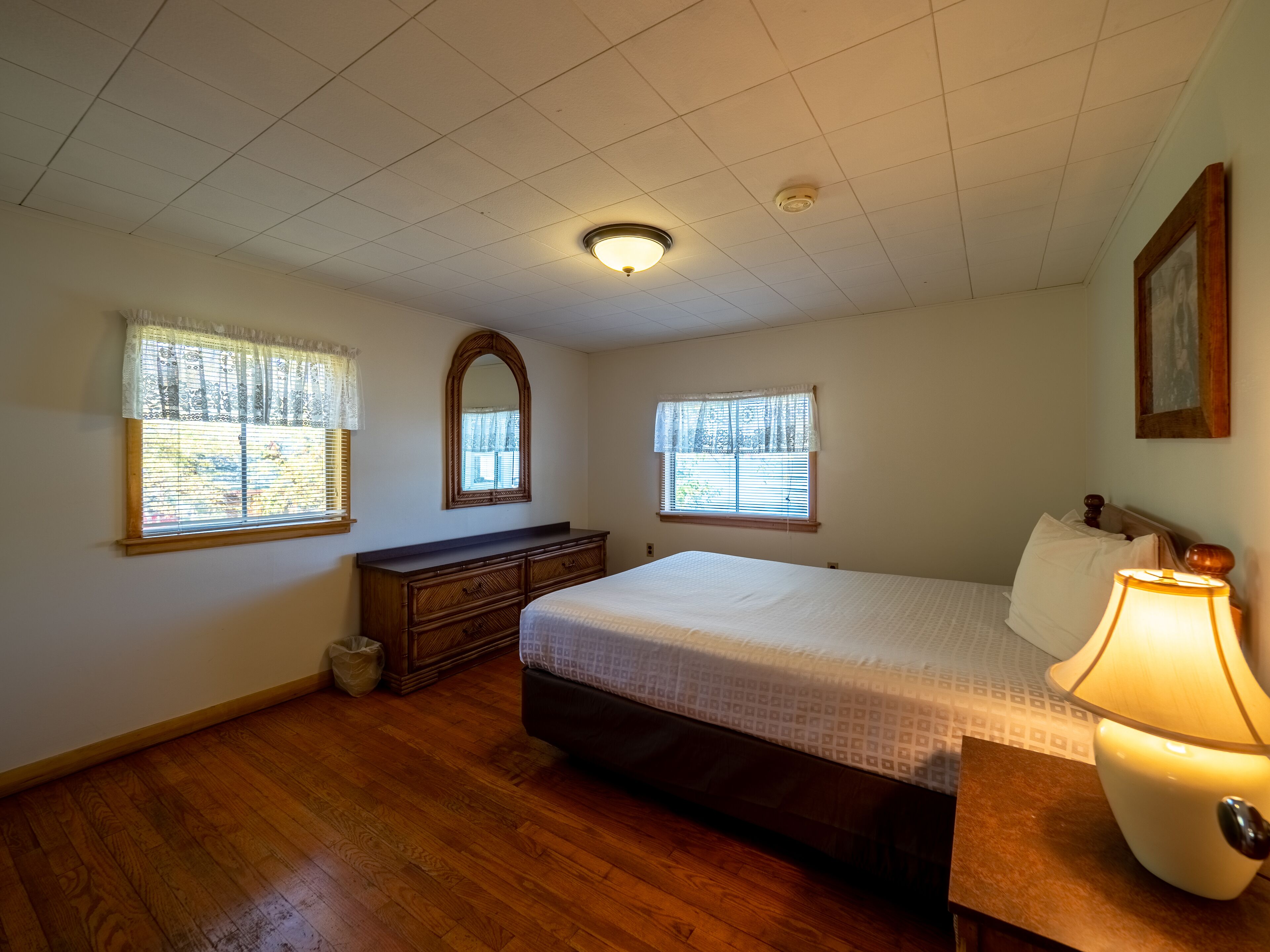 A bedroom with a wooden dresser, a large bed, a mirror, and a window with blinds.