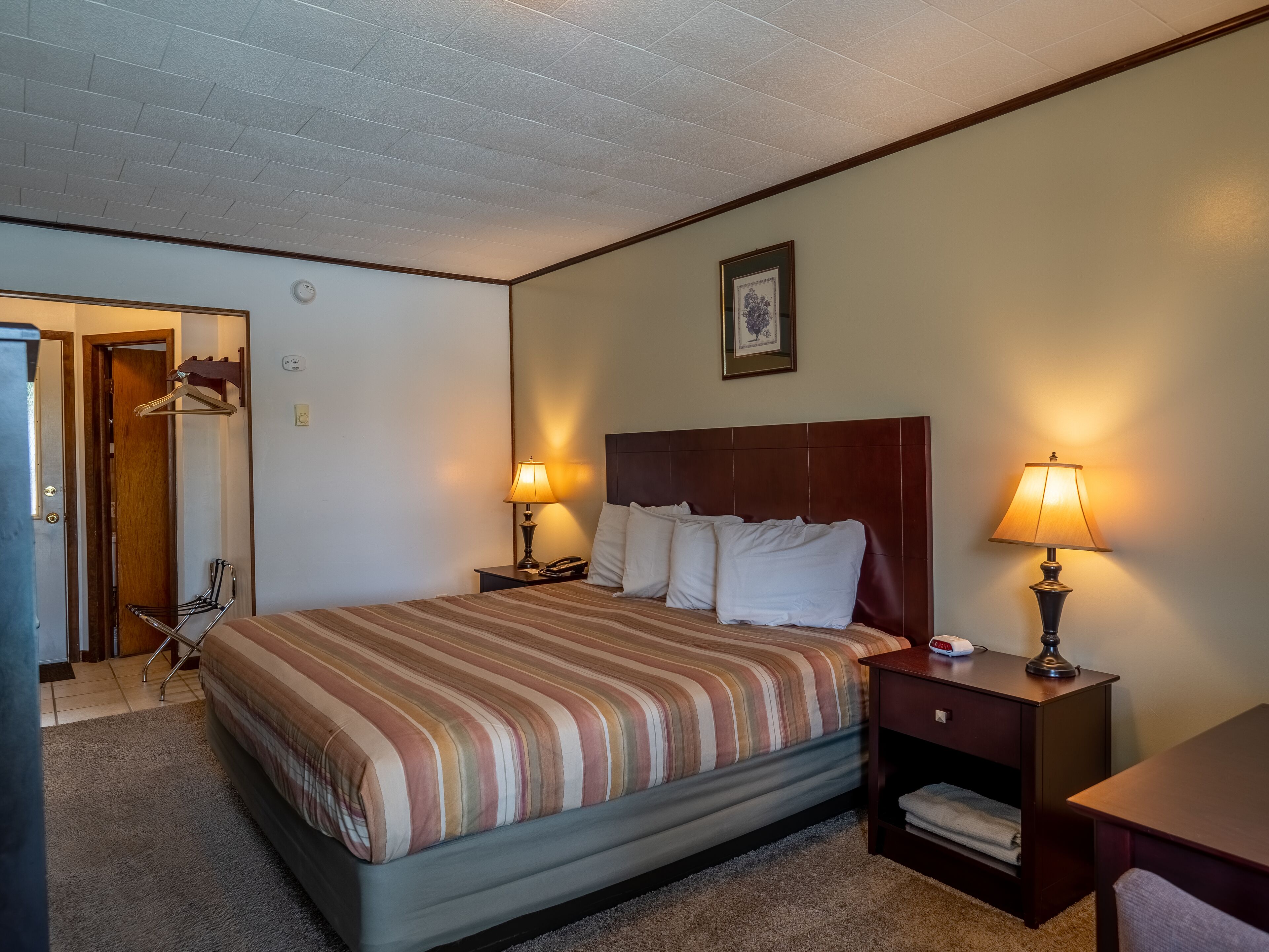 Habitación de hotel con cama, dos mesitas de noche con lámparas, cabecero de madera, un cuadro enmarcado en la pared y una puerta que da a un baño.