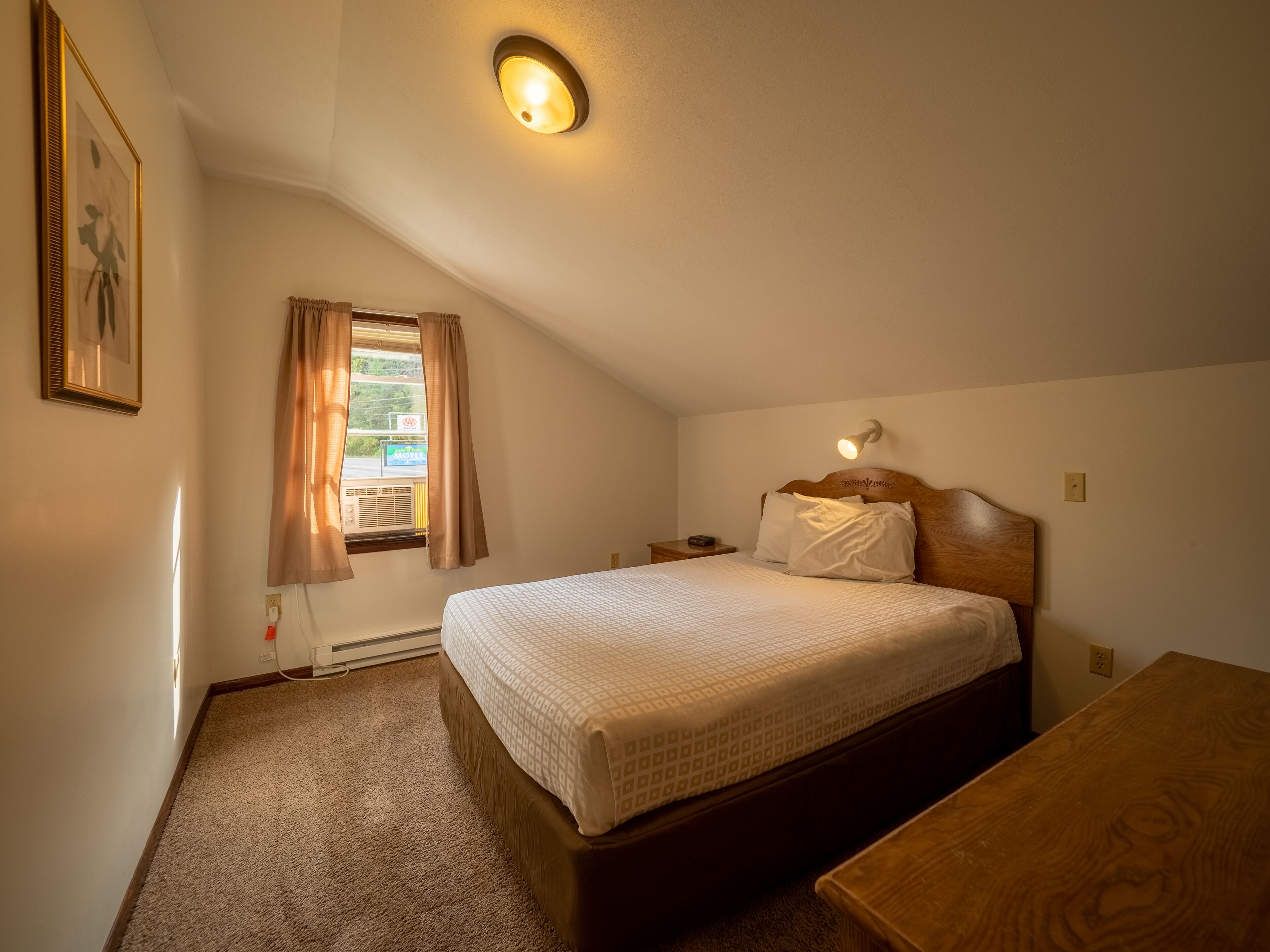 Un dormitorio con cama, mesita de noche, banco de madera, lámpara de pared y ventana con cortinas.