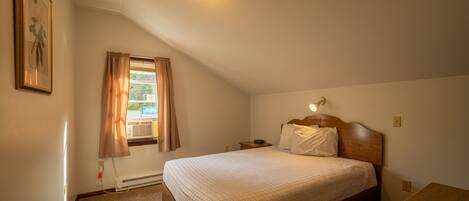 Un dormitorio con cama, mesita de noche, banco de madera, lámpara de pared y ventana con cortinas.