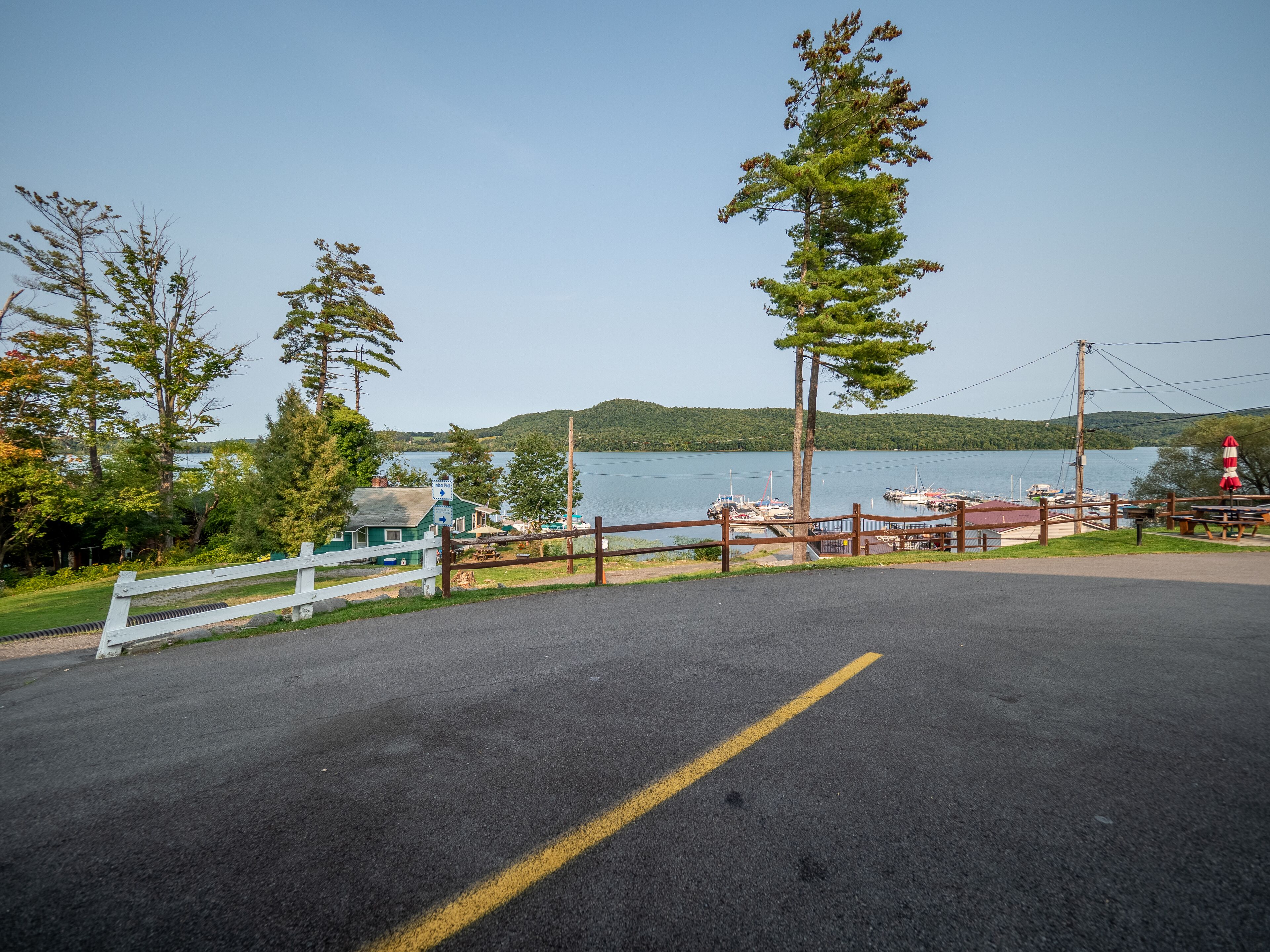 A parking area with a view of a lake, a dock with boats, and a small building with a green roof.