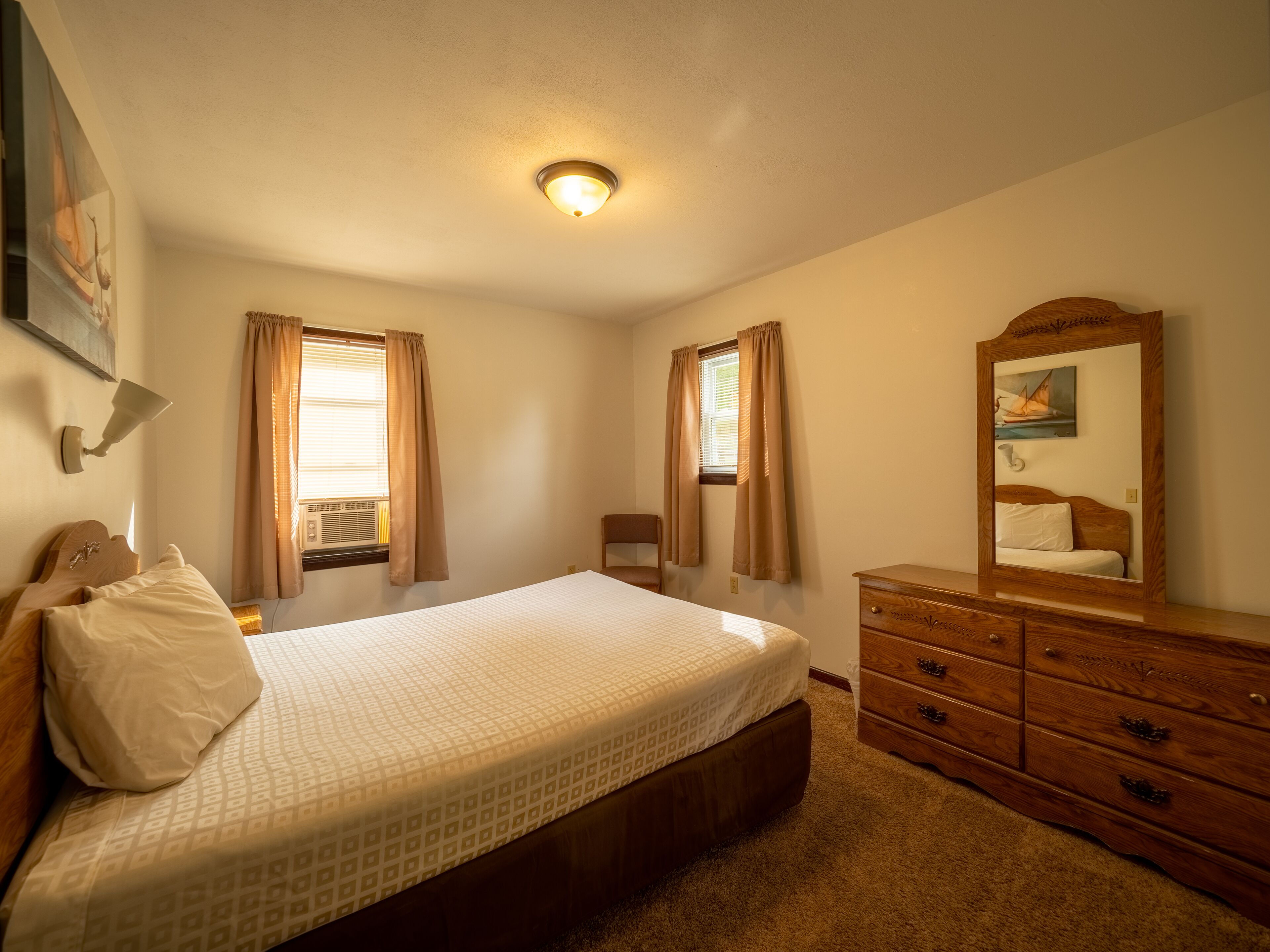 A bedroom with a bed, a dresser, a mirror, and two windows with curtains.