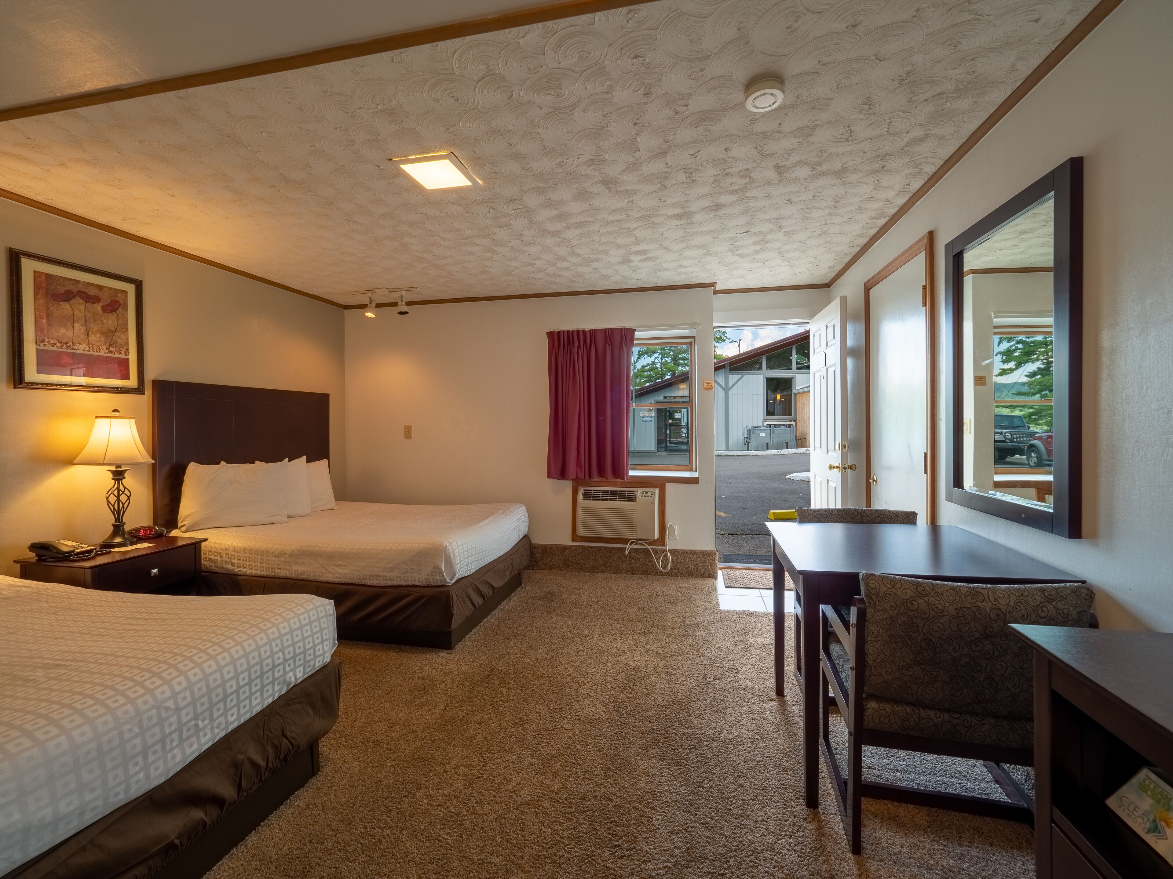A hotel room with two beds, a desk, a chair, a lamp, and a window with a view of the outside.