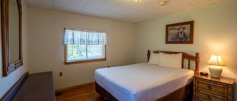A bedroom with a bed, a nightstand, a dresser, a window with blinds, a ceiling light, and a wall-mounted picture.