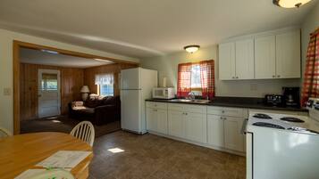A compact kitchen with white cabinets, a refrigerator, a microwave, and a stove. There is a dining table with chairs and a window with curtains.