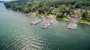 An aerial view of a lakeside village with boats docked along the shore, surrounded by lush greenery and residential buildings. - Lake 'N Pines Motel (Cooperstown)