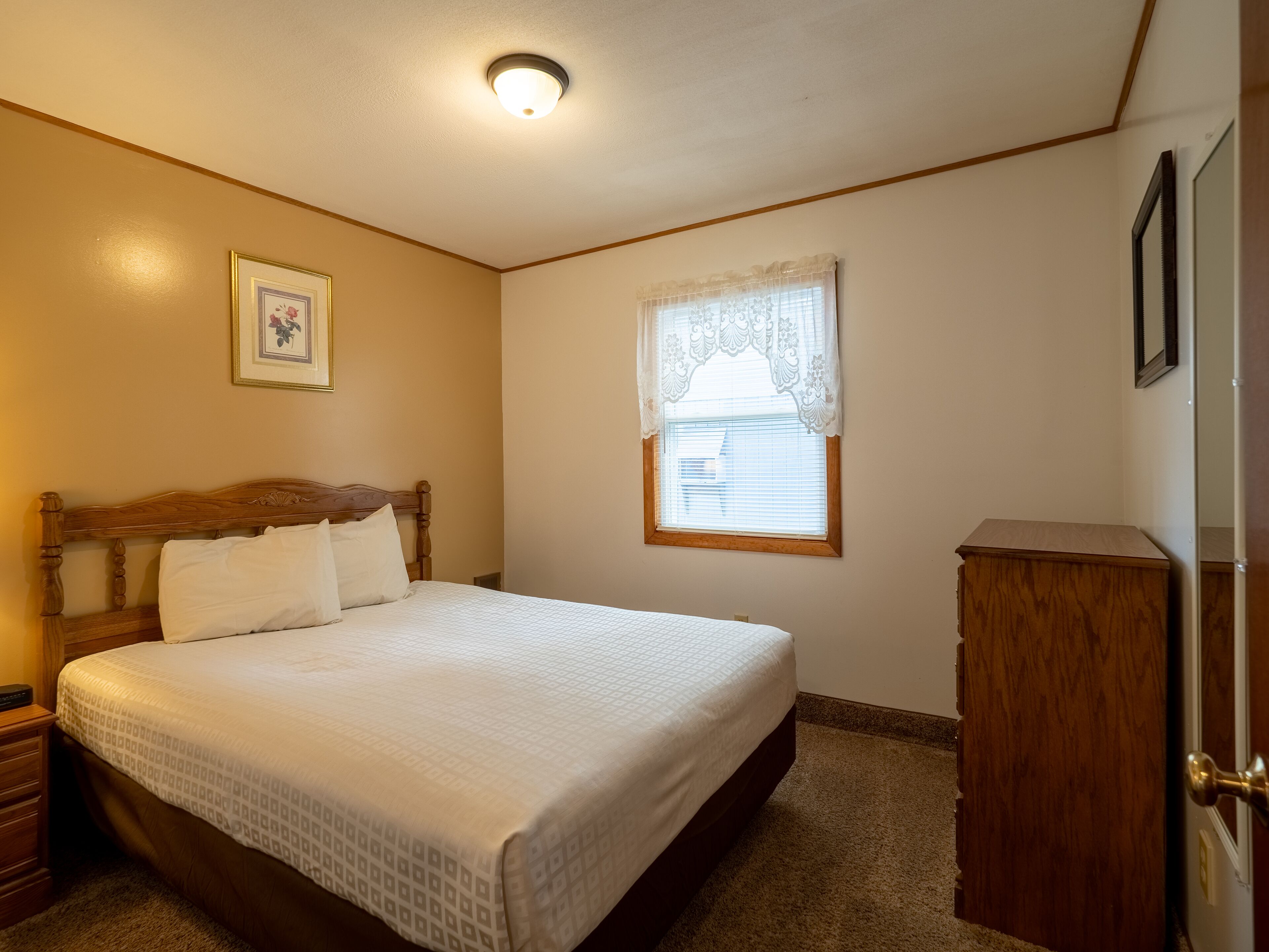 A bedroom with a wooden bed, a nightstand, a dresser, a window with lace curtains, and a ceiling light.