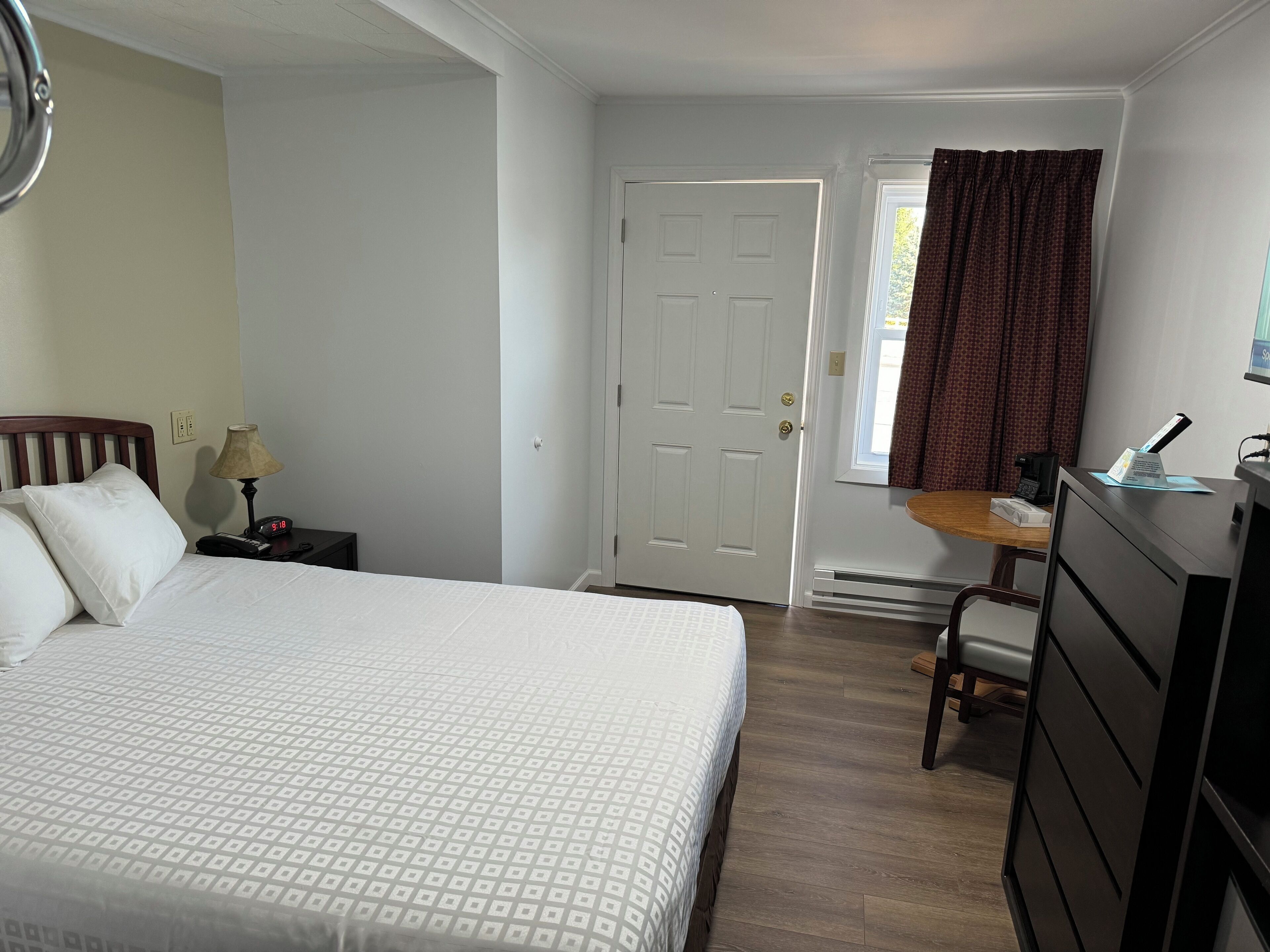 Camera d'albergo con un letto, una scrivania, una sedia, un comò, una lampada, una sveglia, una porta e una finestra con tende.