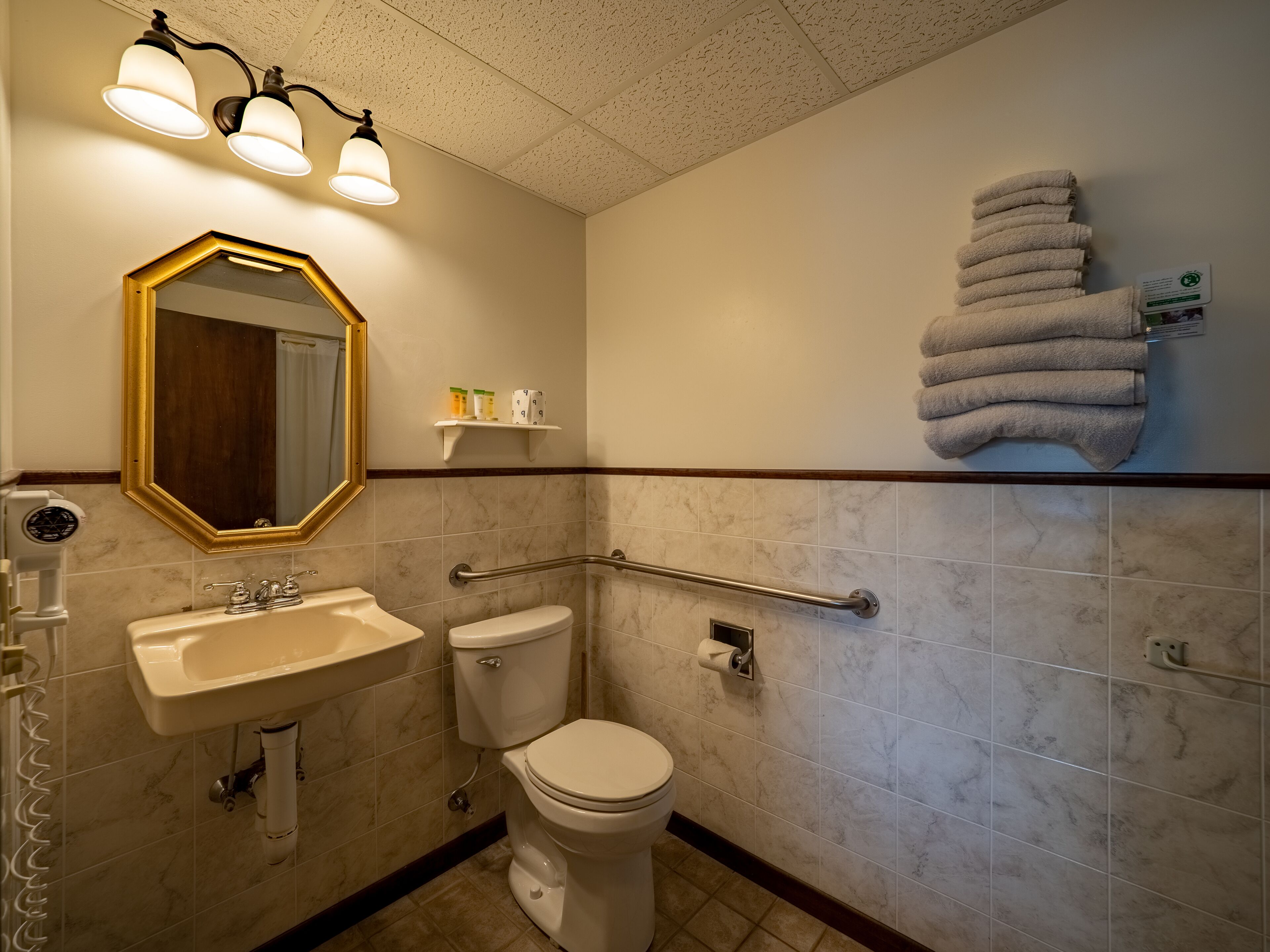 A bathroom with a sink, toilet, and a mirror. There are towels stacked on the wall and a handrail.